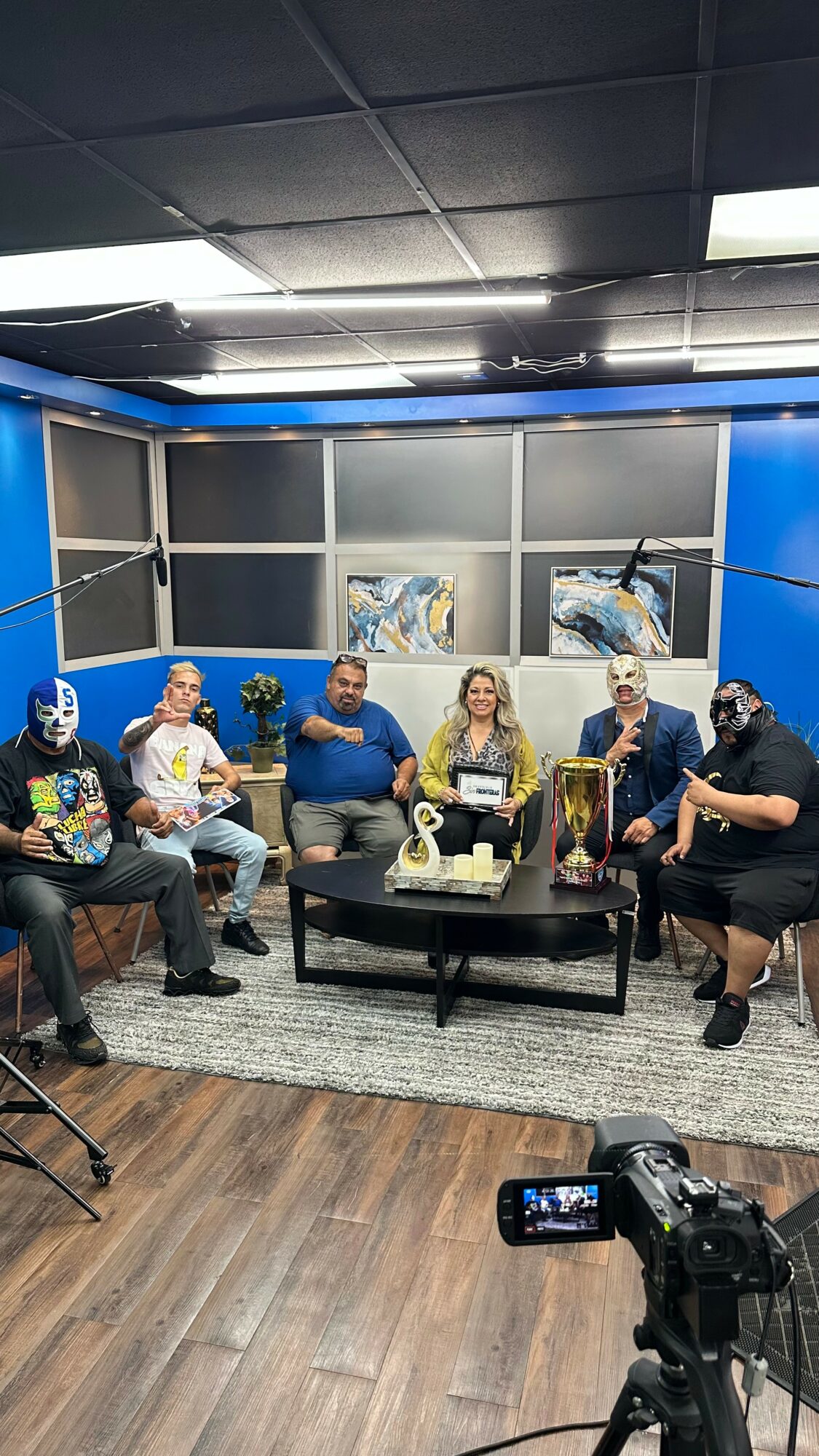 Six people sitting on a sofa and chairs in a studio with blue walls, a coffee table, and artwork, facing camera.