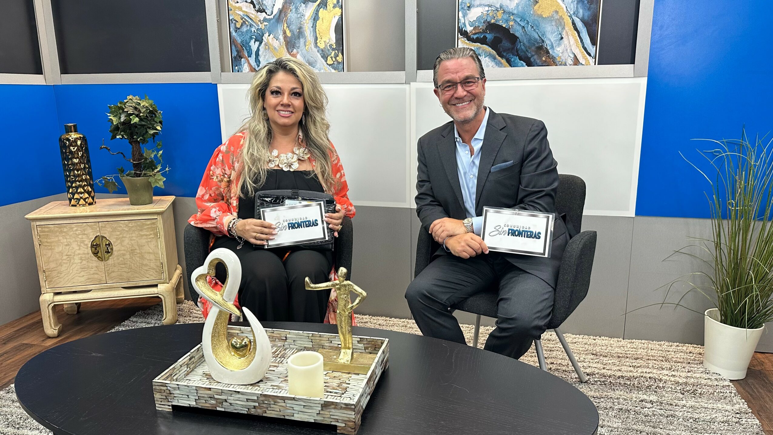 Two people sitting on chairs holding framed certificates, with decorative items and plants in the background.