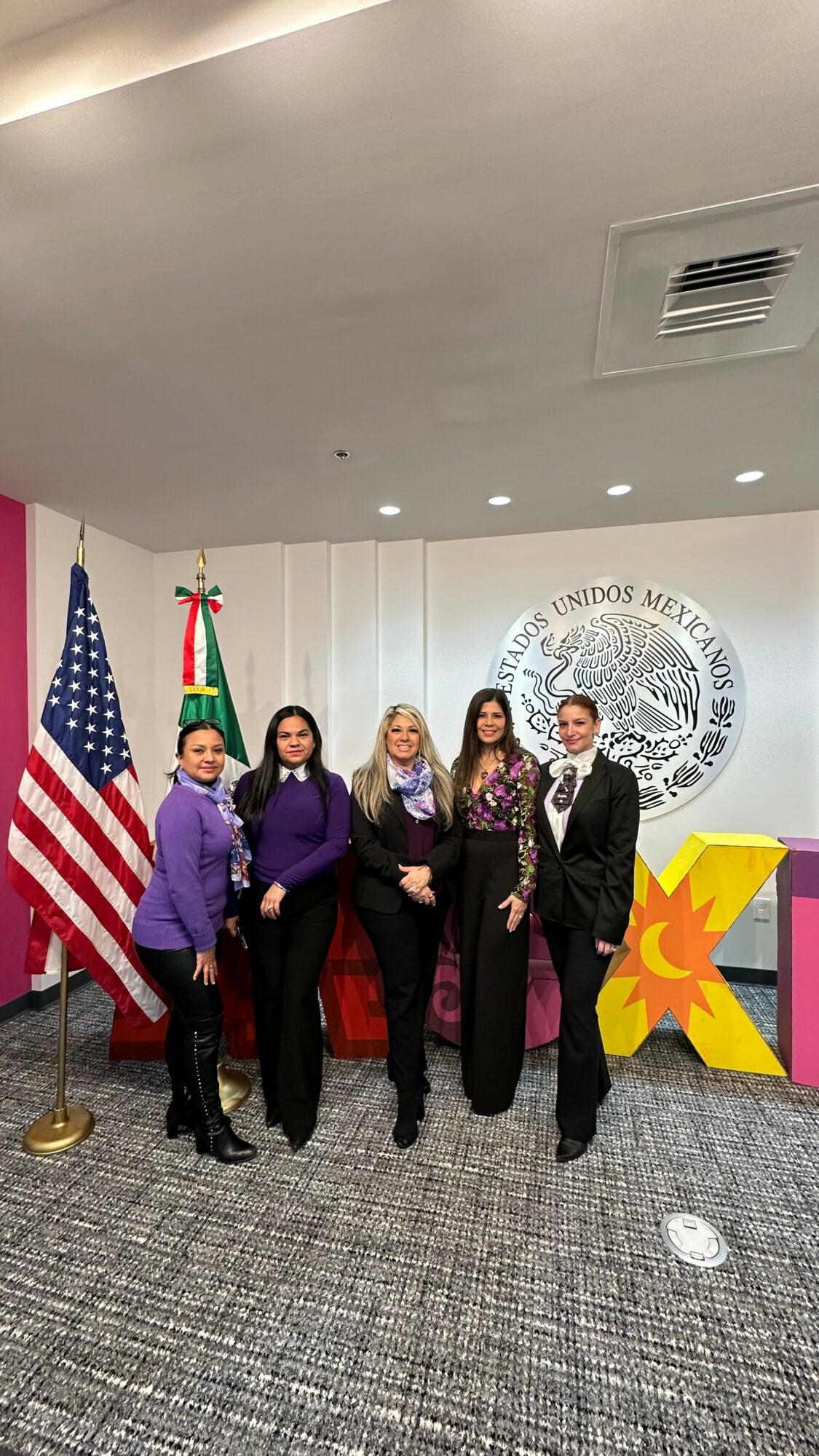 Five women standing in front of a Mexican government seal, with an American flag and Mexican flag behind them, in a formal setting.