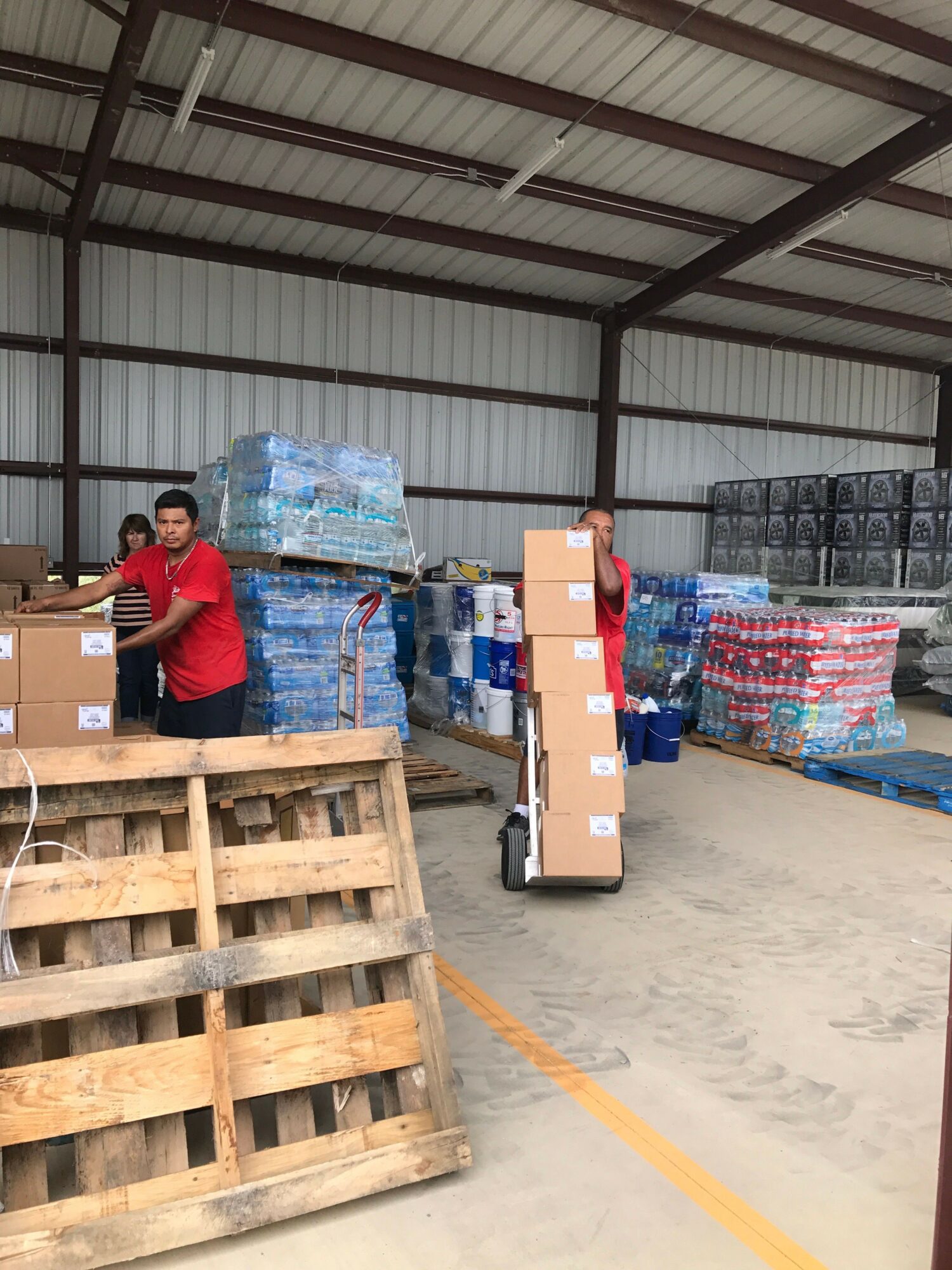 Warehouse interior with stacked boxes, pallets, and a person in red shirt handling boxes.
