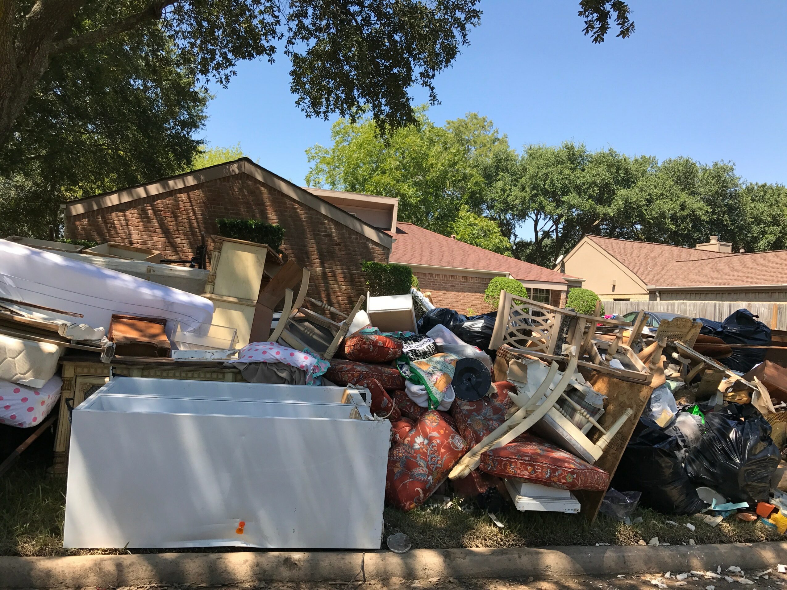 Piles of discarded furniture, bags, and debris outdoors near houses and trees, with a clear blue sky.