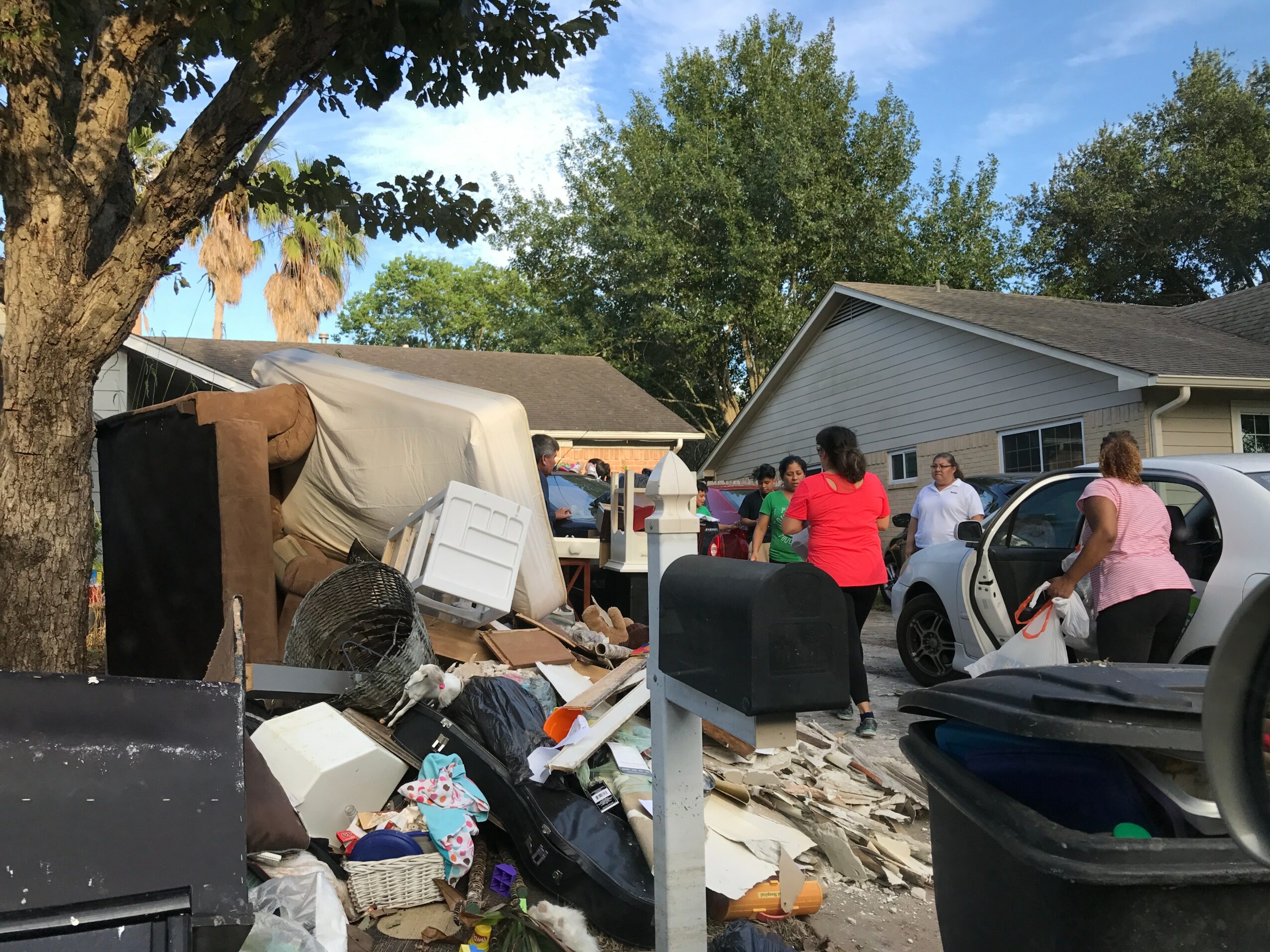 People gather outside a house with debris and damaged items on the ground, under a tree and blue sky.