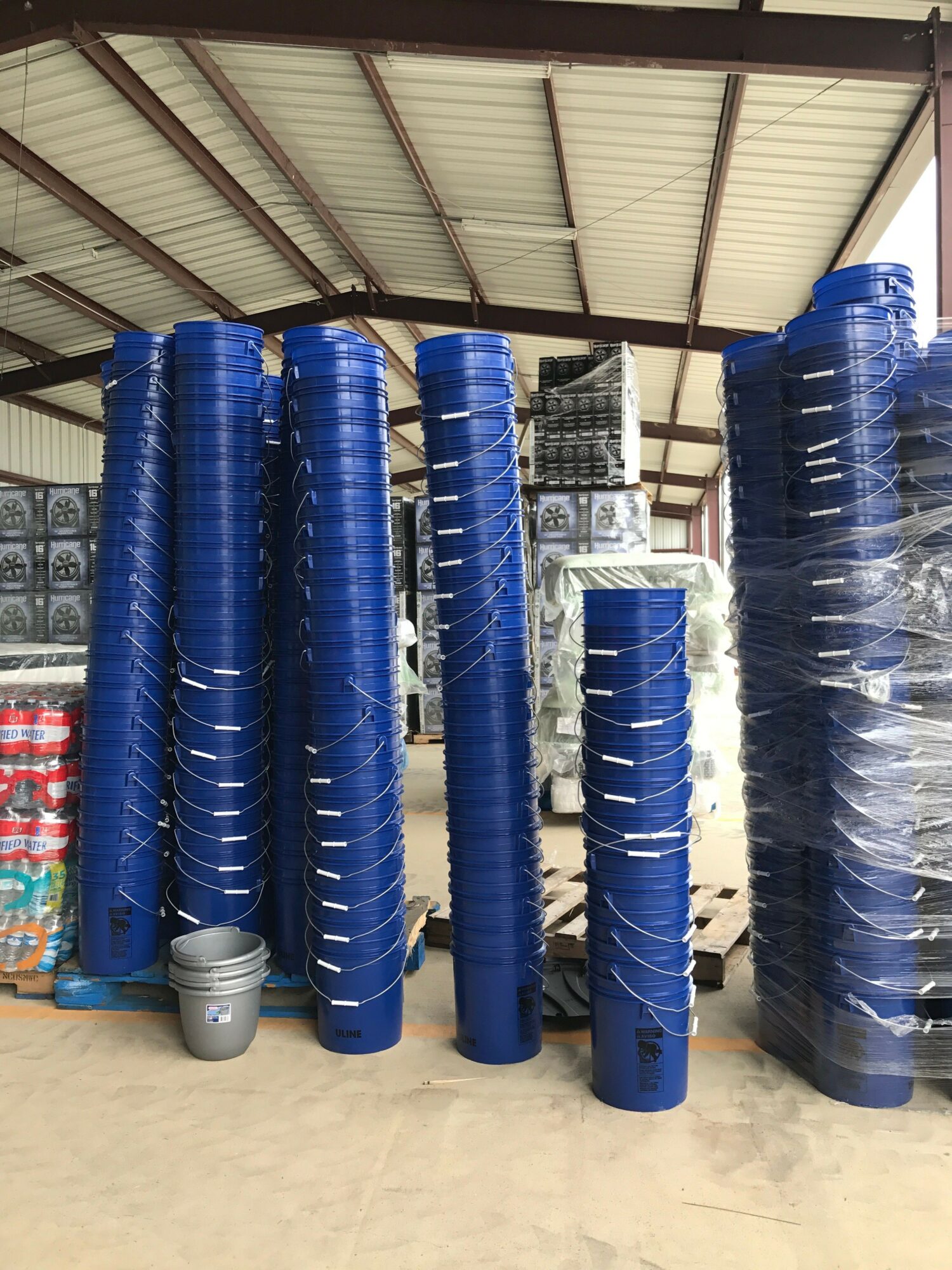 Rows of blue water filter cartridges stored in a warehouse, with some wrapped in plastic, and a metal roof overhead.