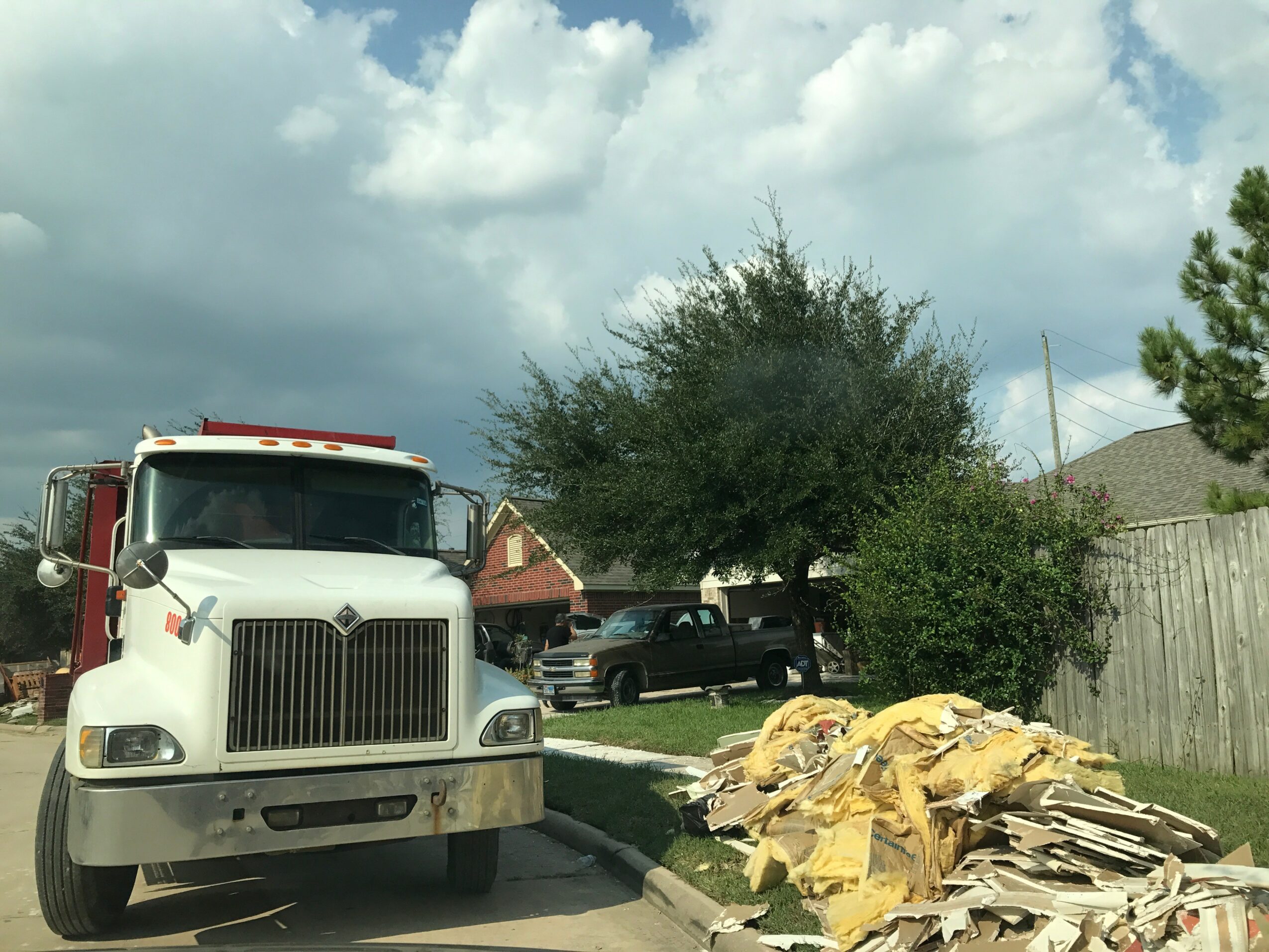 White truck parked on street with trees and houses in background, pile of cardboard and paper on sidewalk, cloudy sky.