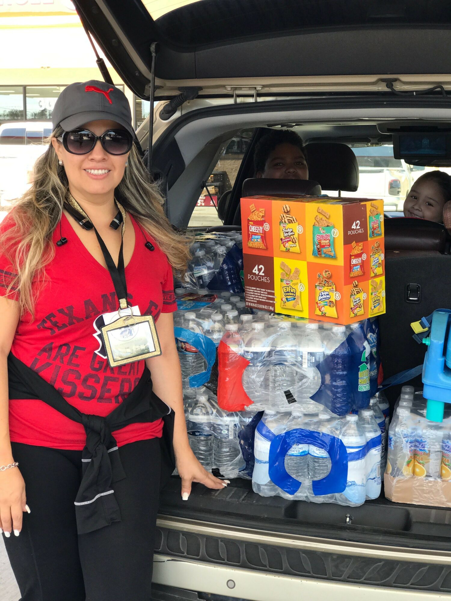 Woman in red shirt and sunglasses sitting in car trunk filled with water bottles and snacks, holding a tablet.
