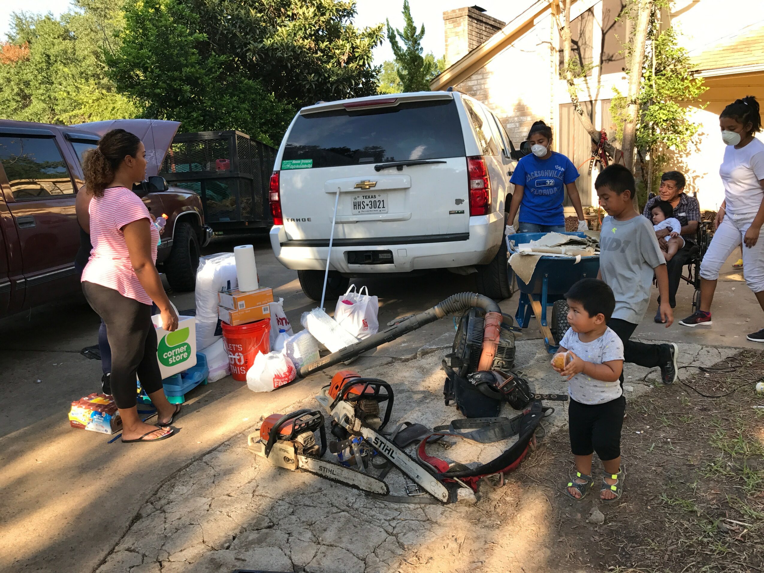 Group of children and adults outdoors near parked cars, with tools and equipment on the ground, on a sunny day.