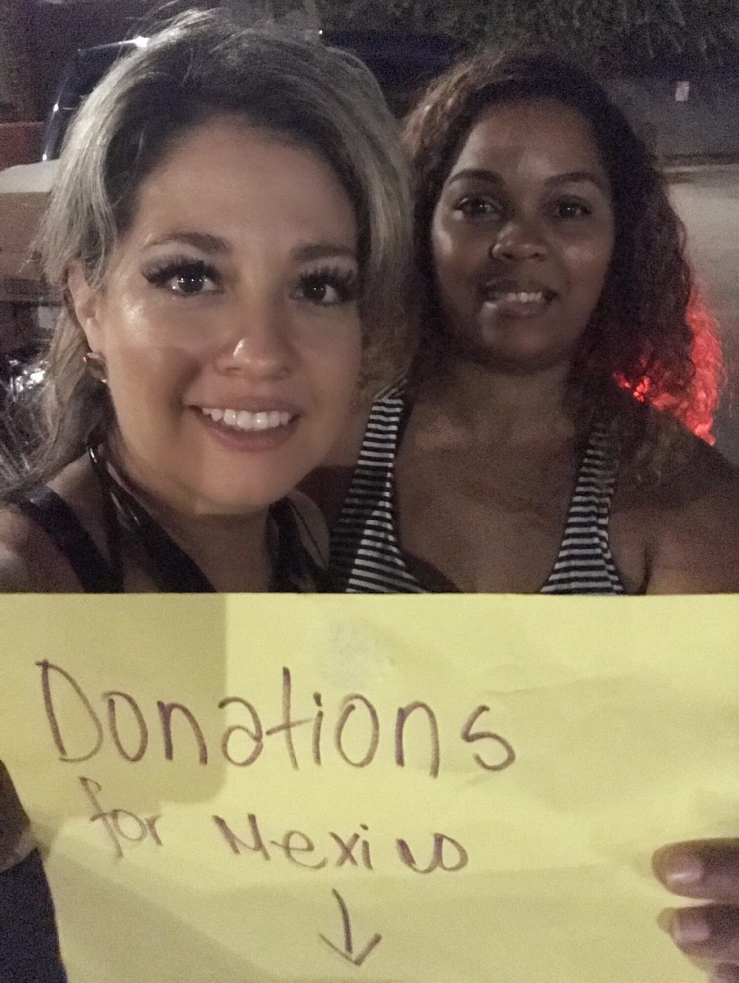 Two women smiling, holding a yellow sign that says 'Donations for Mexico' with an arrow, outdoors at night.