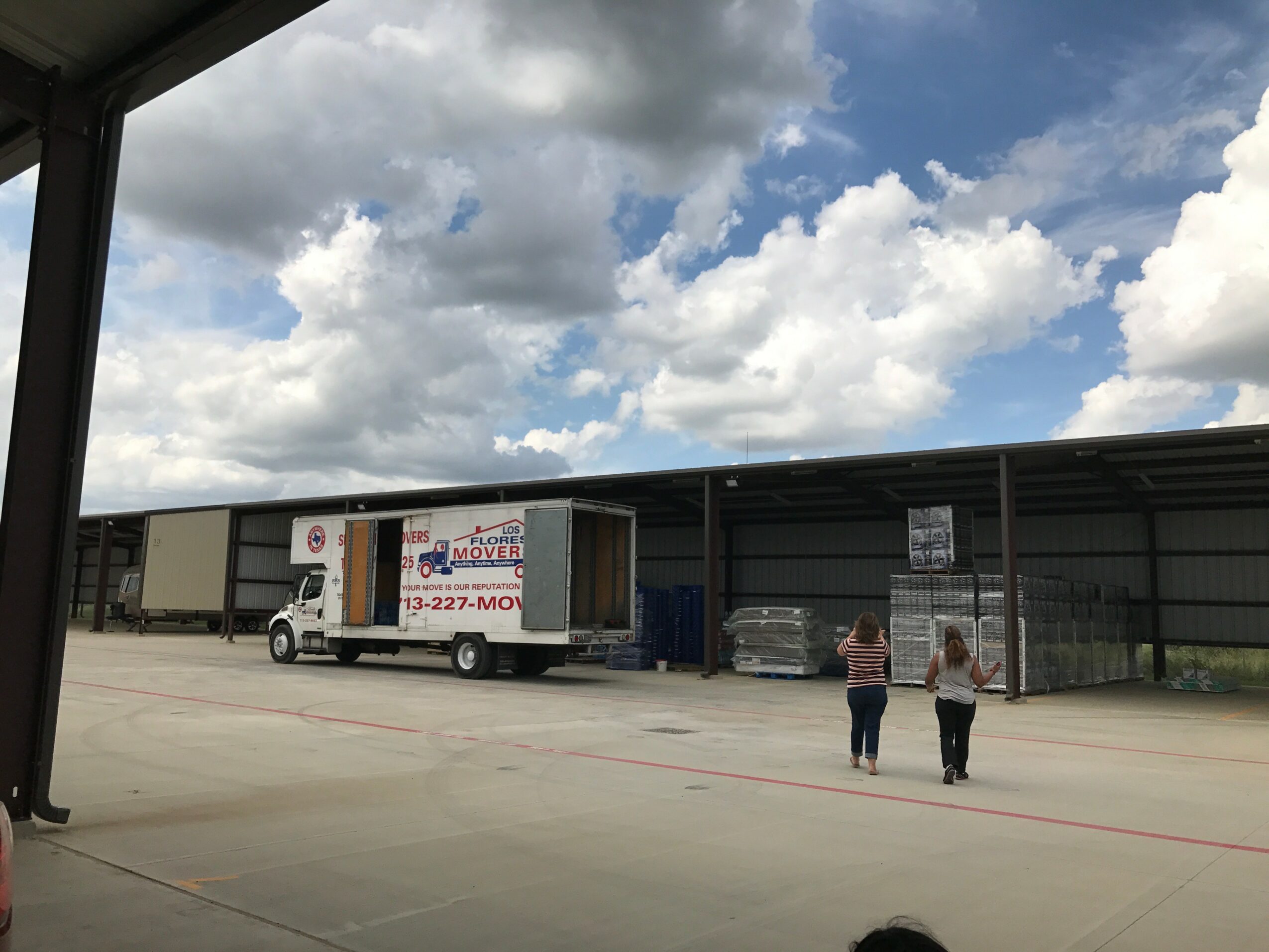 Two children walk on a large open area near a warehouse with trucks and parked cars, under a partly cloudy sky.