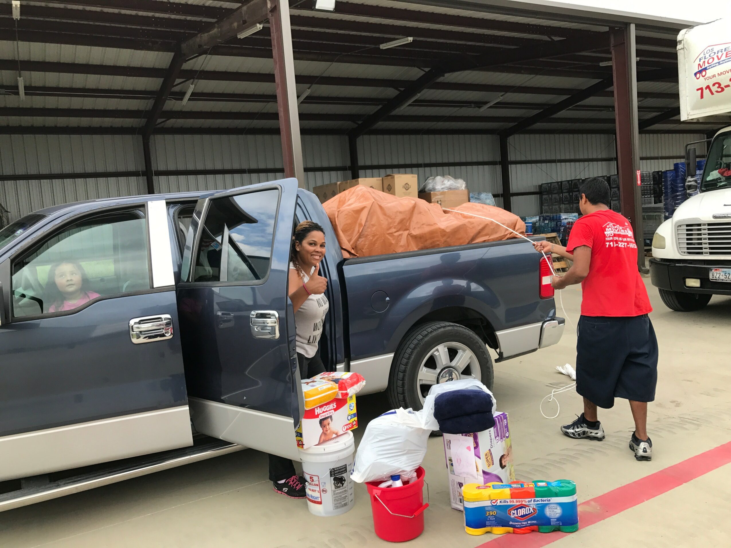 People loading supplies into a pickup truck in a warehouse area, with boxes and bottles nearby.