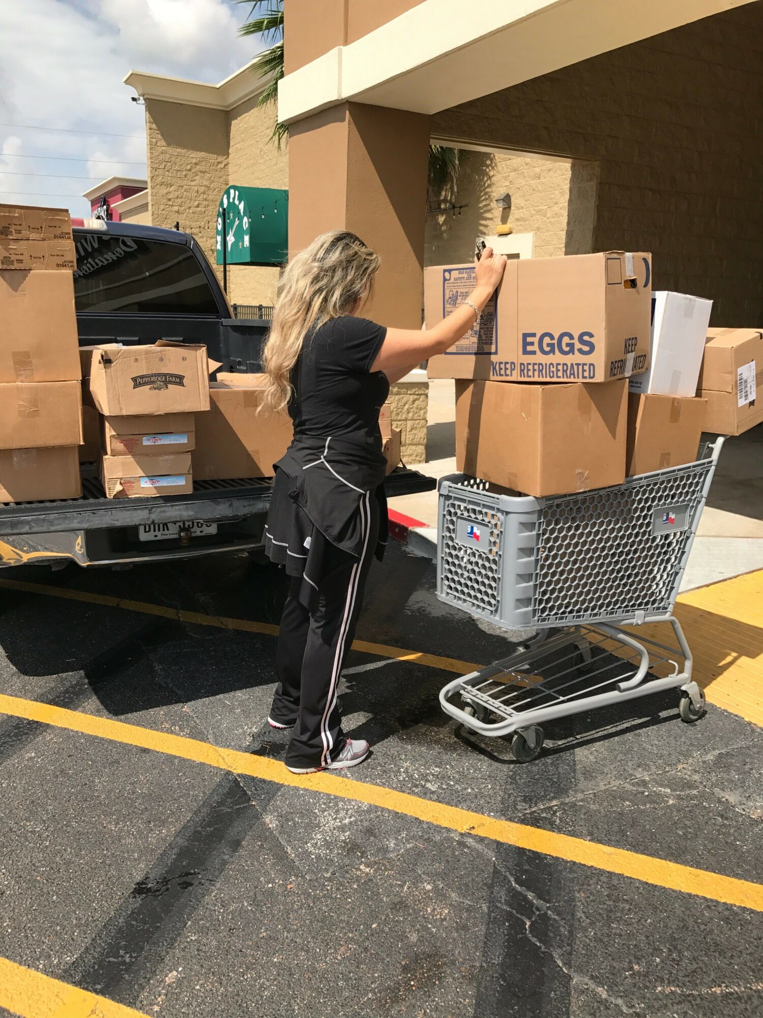 Person loading boxes into a truck outside, with a shopping cart nearby, on a paved area with yellow lines.