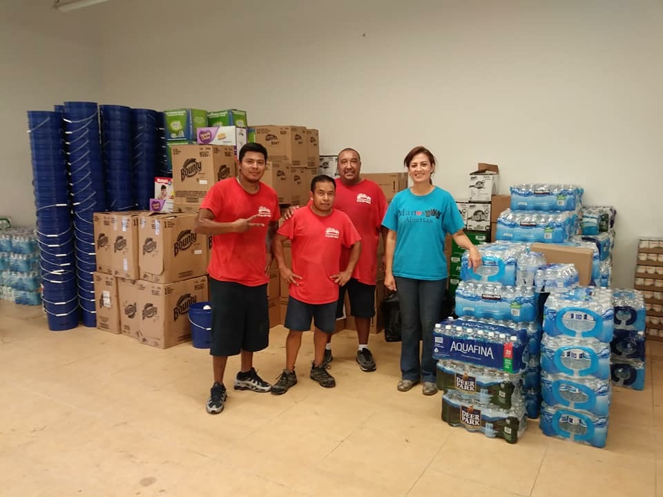 Four people standing in a room with stacked boxes and bottled water, smiling at the camera.