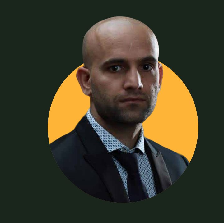Bald man in a suit and patterned shirt against a yellow circle background, with a dark outer background.