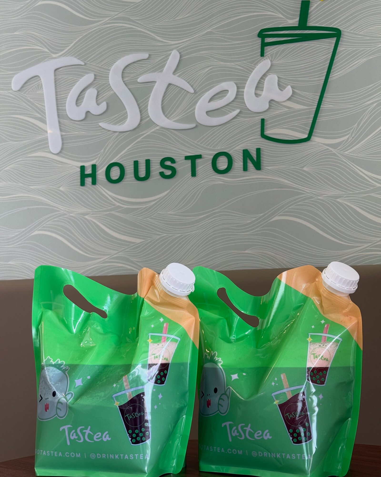 Two green drink pouches with white caps and straws, in front of a wall with 'Tasted Houston' logo and a drink icon.