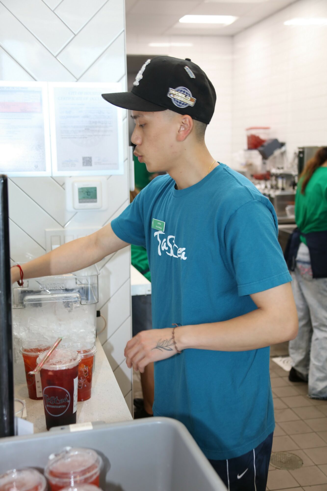 Young man in blue shirt and black cap at a counter with drinks, in a fast-food restaurant, facing left.