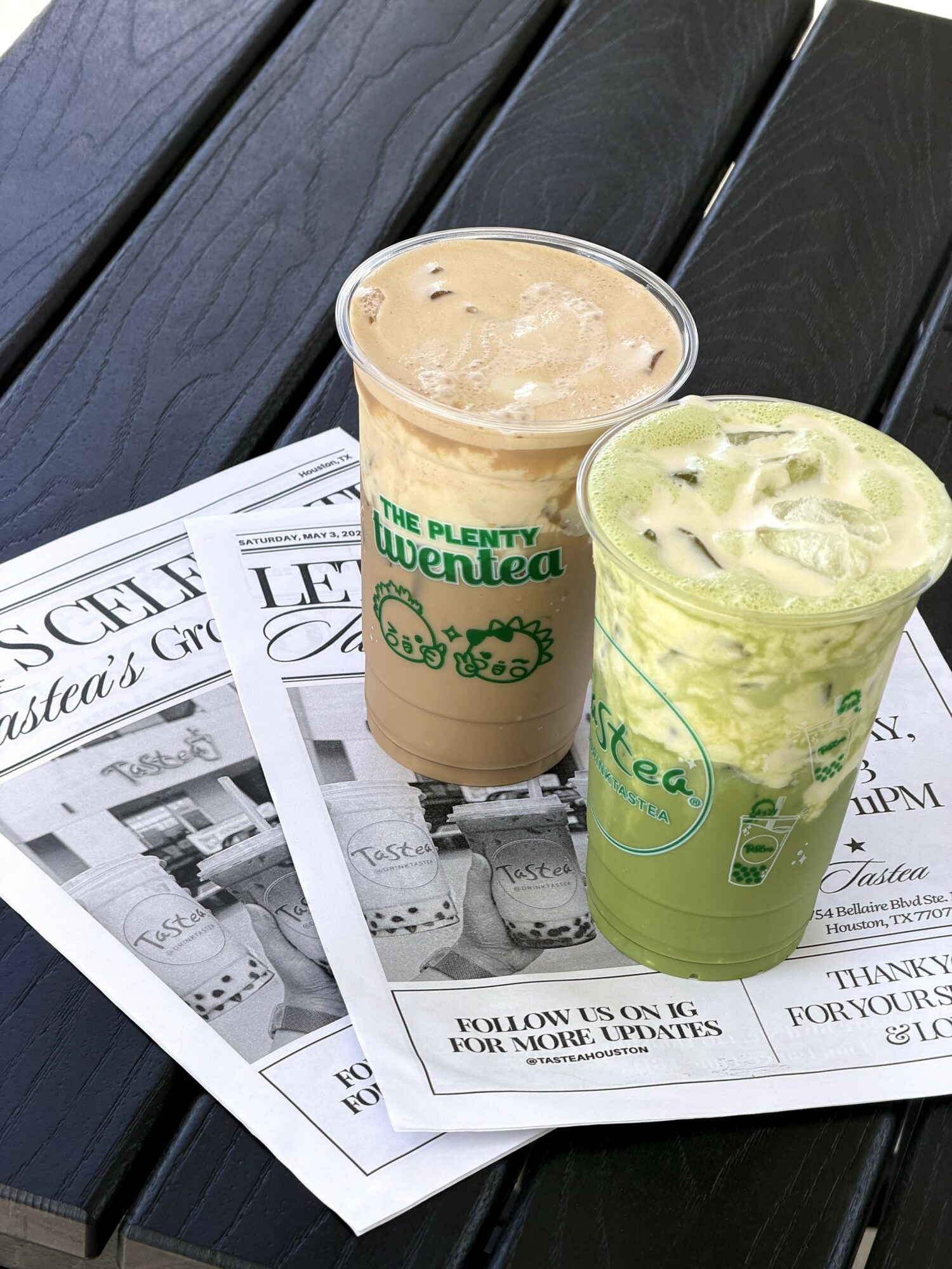 Two iced beverages in plastic cups with green logos on a black wooden surface, placed on newspapers.