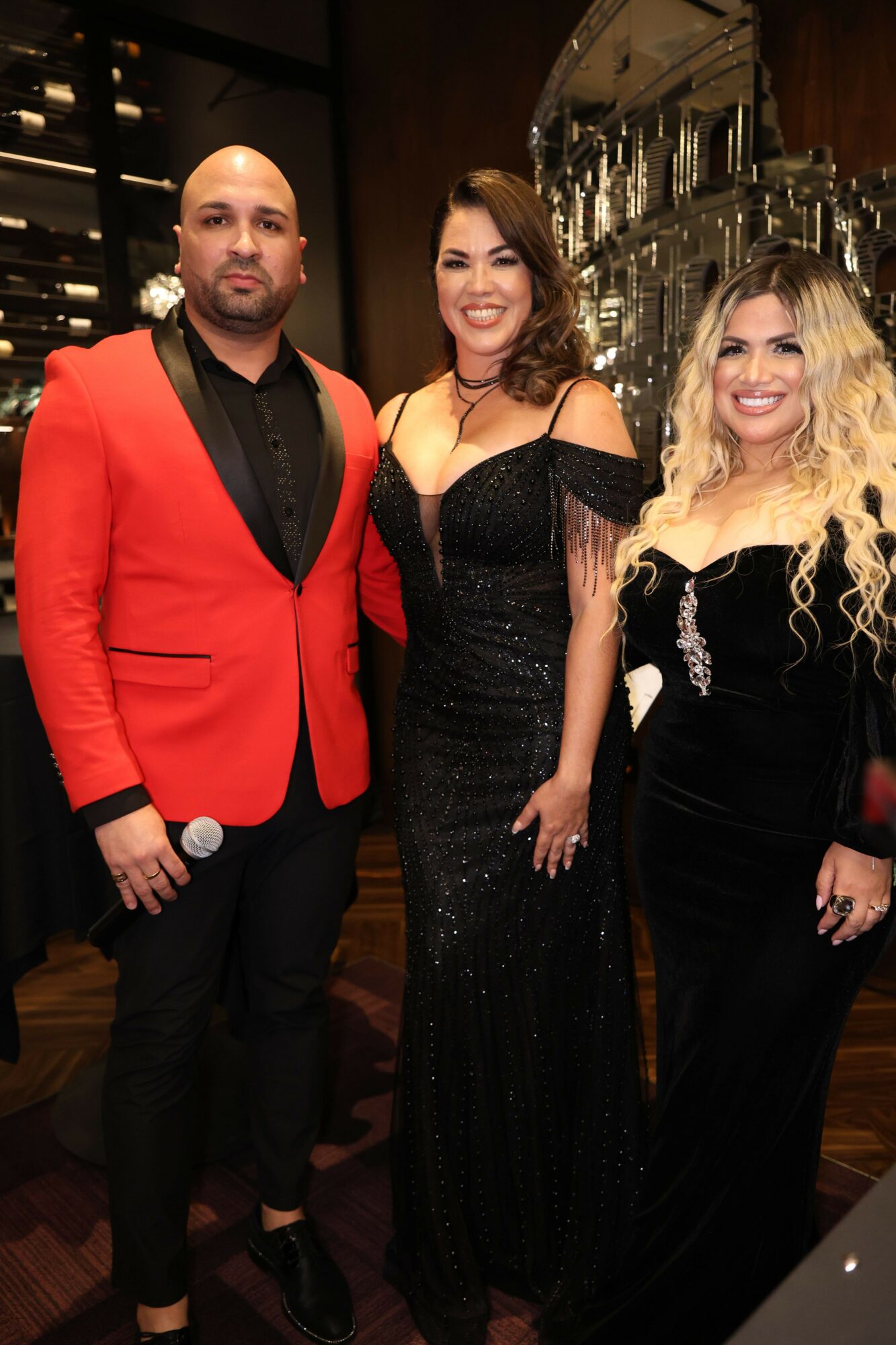 Two women and one man dressed in formal attire at a social event, smiling and posing indoors with a bar in the background.