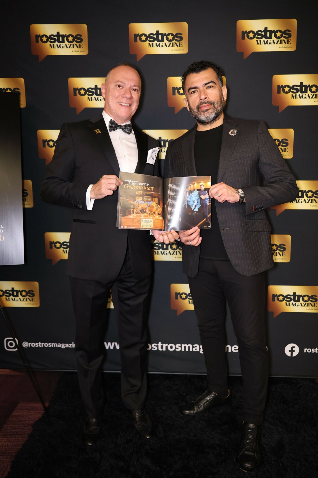 Two men in suits holding a framed certificate and a book, standing in front of a backdrop with logos.