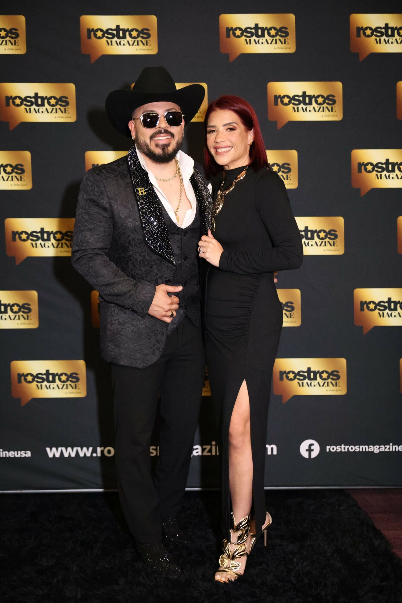 Two women posing at an event with a backdrop featuring logos, one in a black dress and the other in a coat and sunglasses.