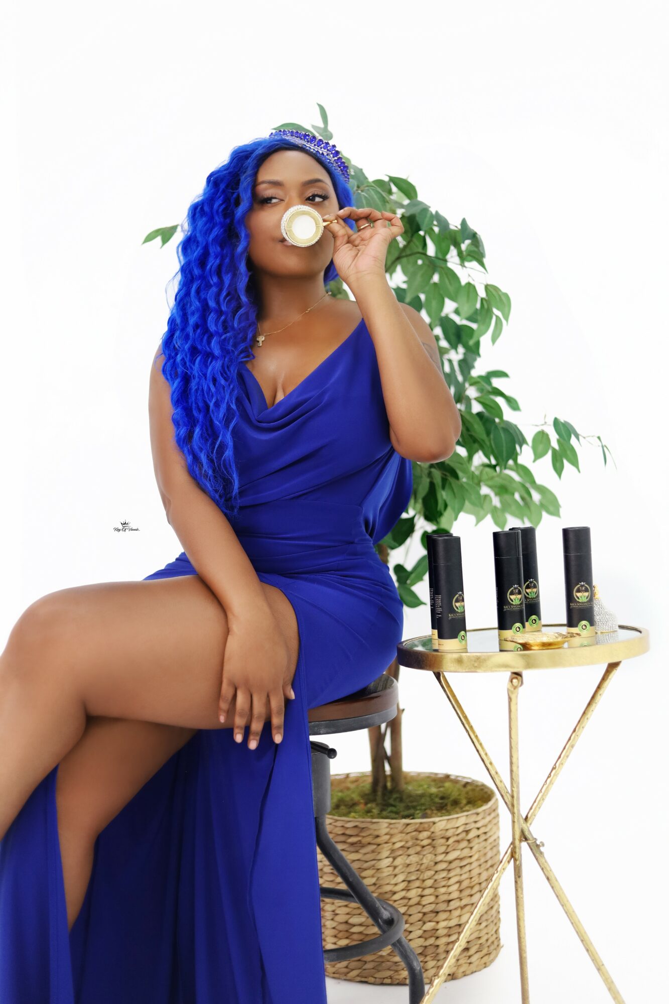 Woman with blue curly hair in a blue dress sitting on a chair, holding a spoon near her eye, with a plant and candles nearby.