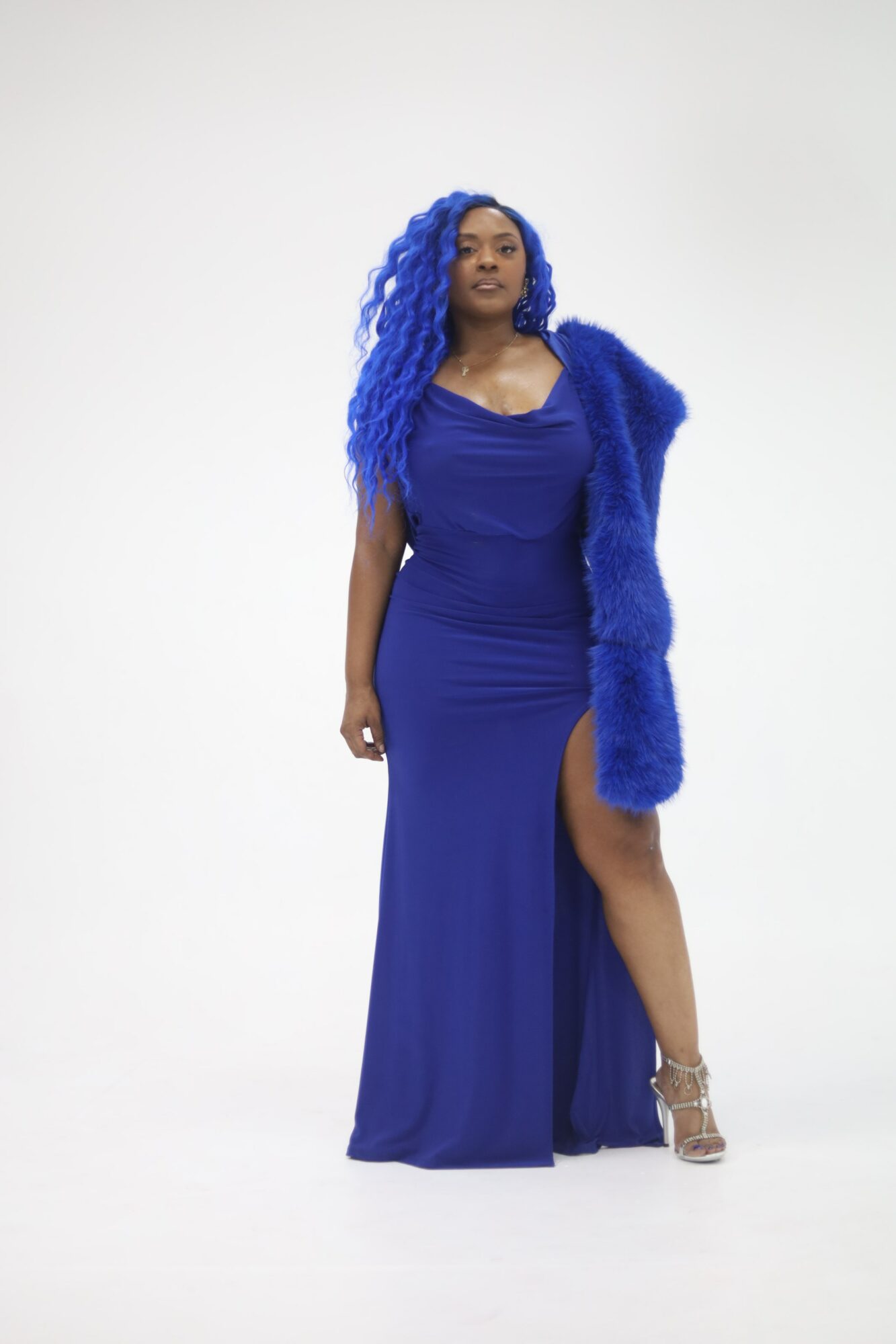 Woman in a long blue dress with a slit, wearing a matching blue fur stole and silver heels, standing against a plain white background.