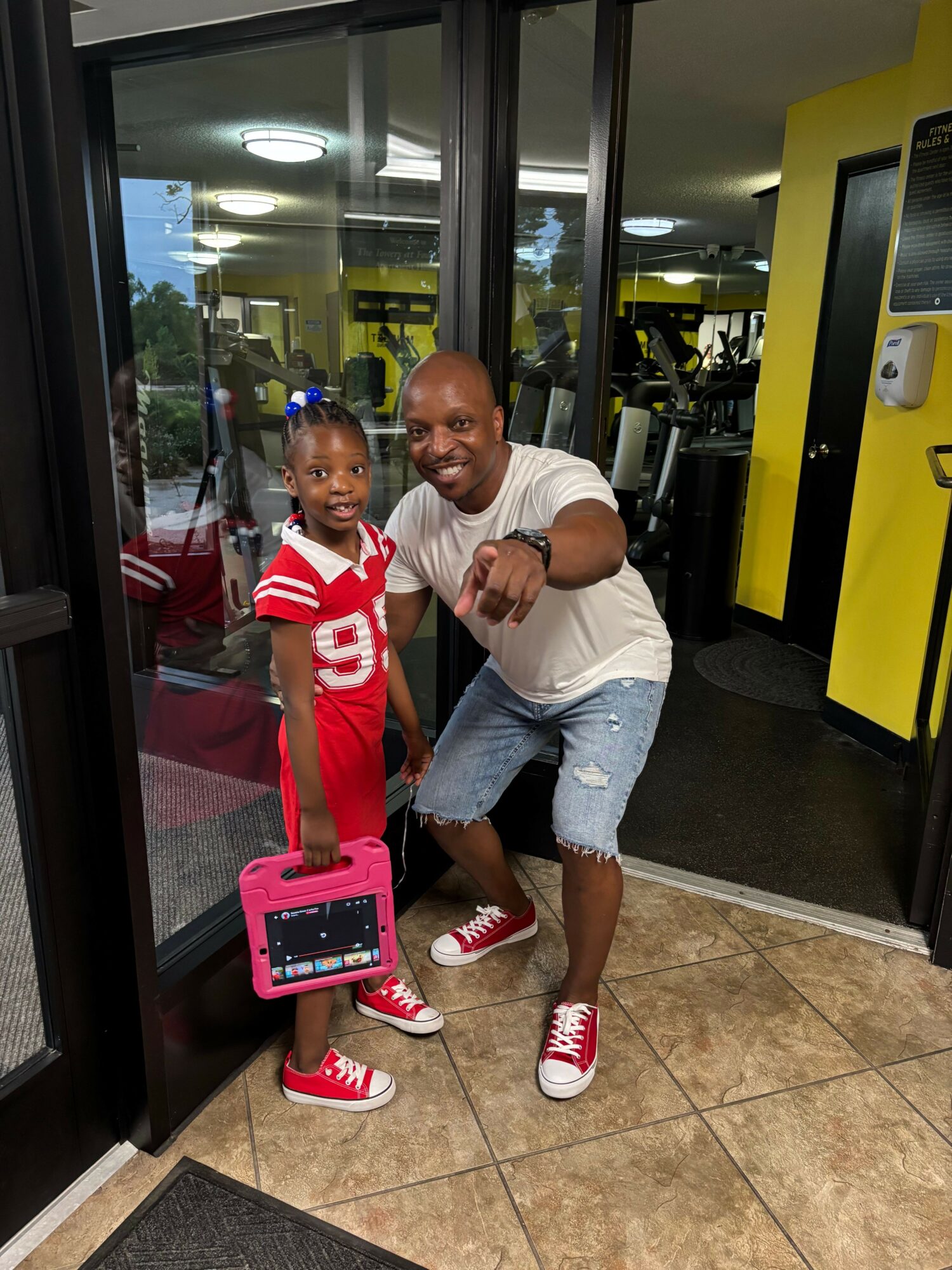 A man and a young girl are taking a selfie in front of a mirror, both smiling and pointing at the camera.