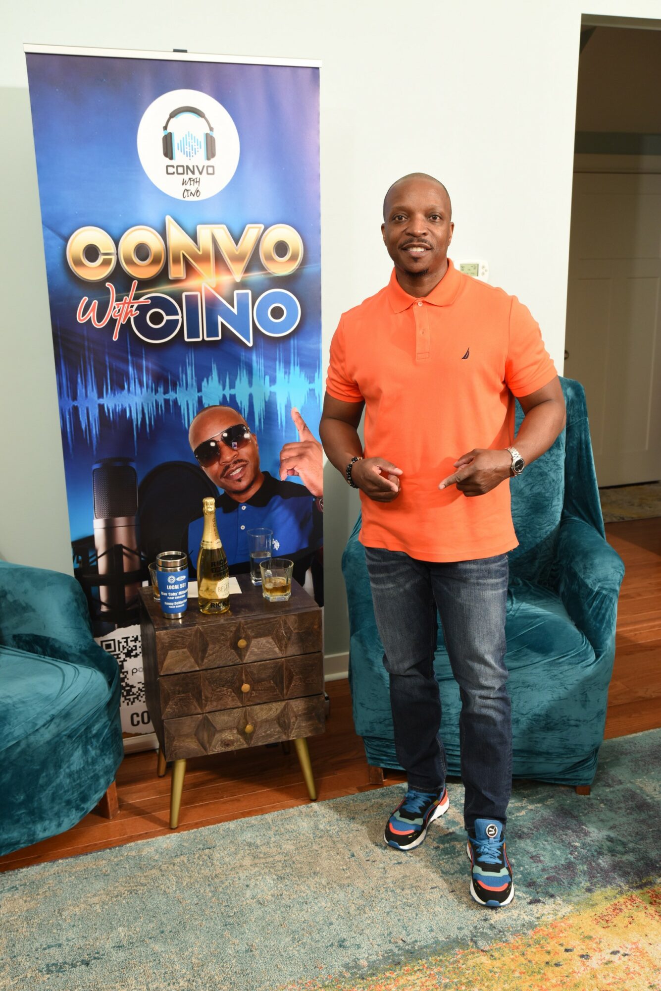 Man in orange shirt and jeans standing next to a promotional banner, holding a phone, with chairs nearby.