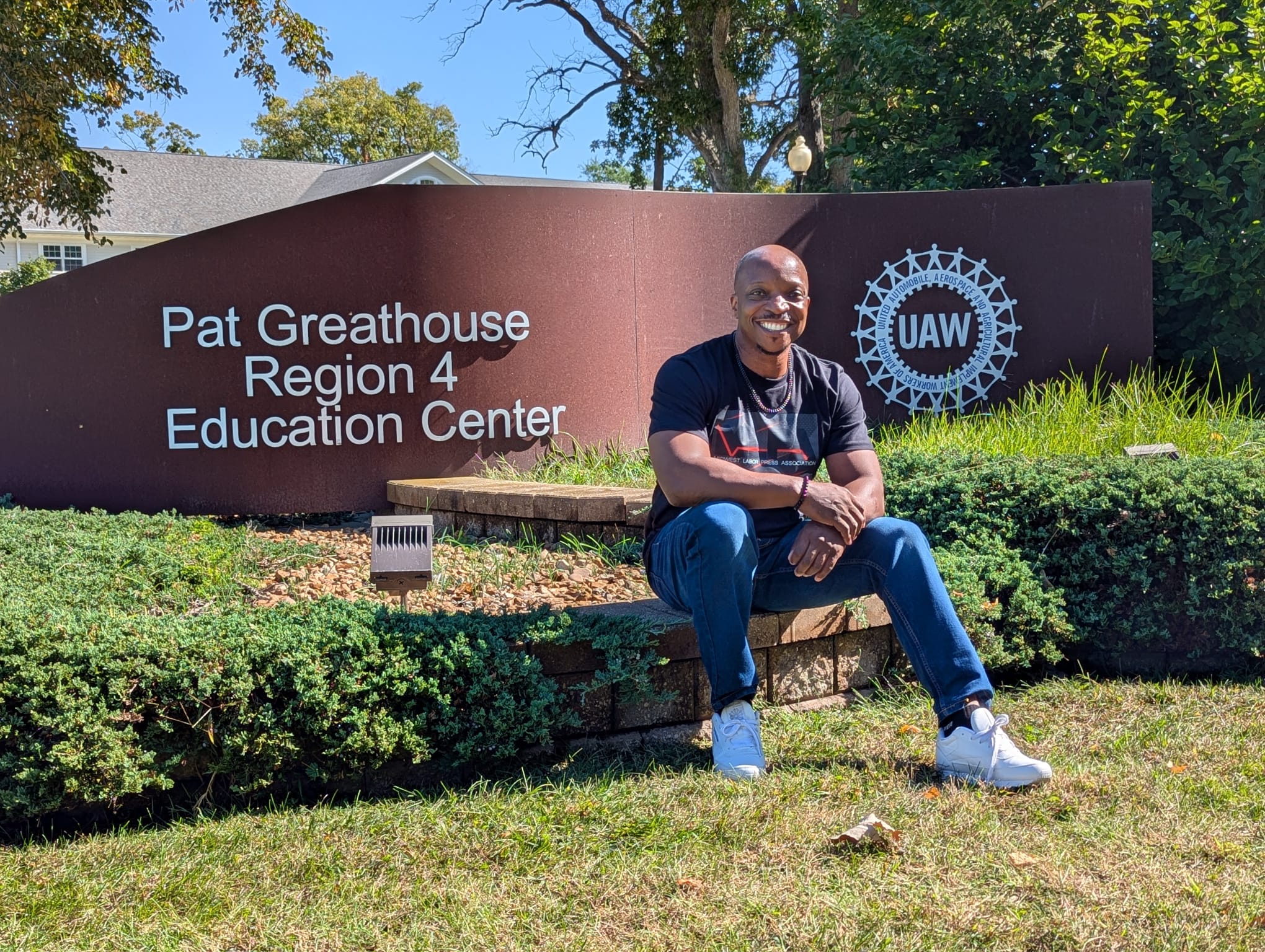 Person sitting on a low brick wall in front of a sign for Pat Greathouse Region 4 Education Center, outdoors with trees and blue sky.