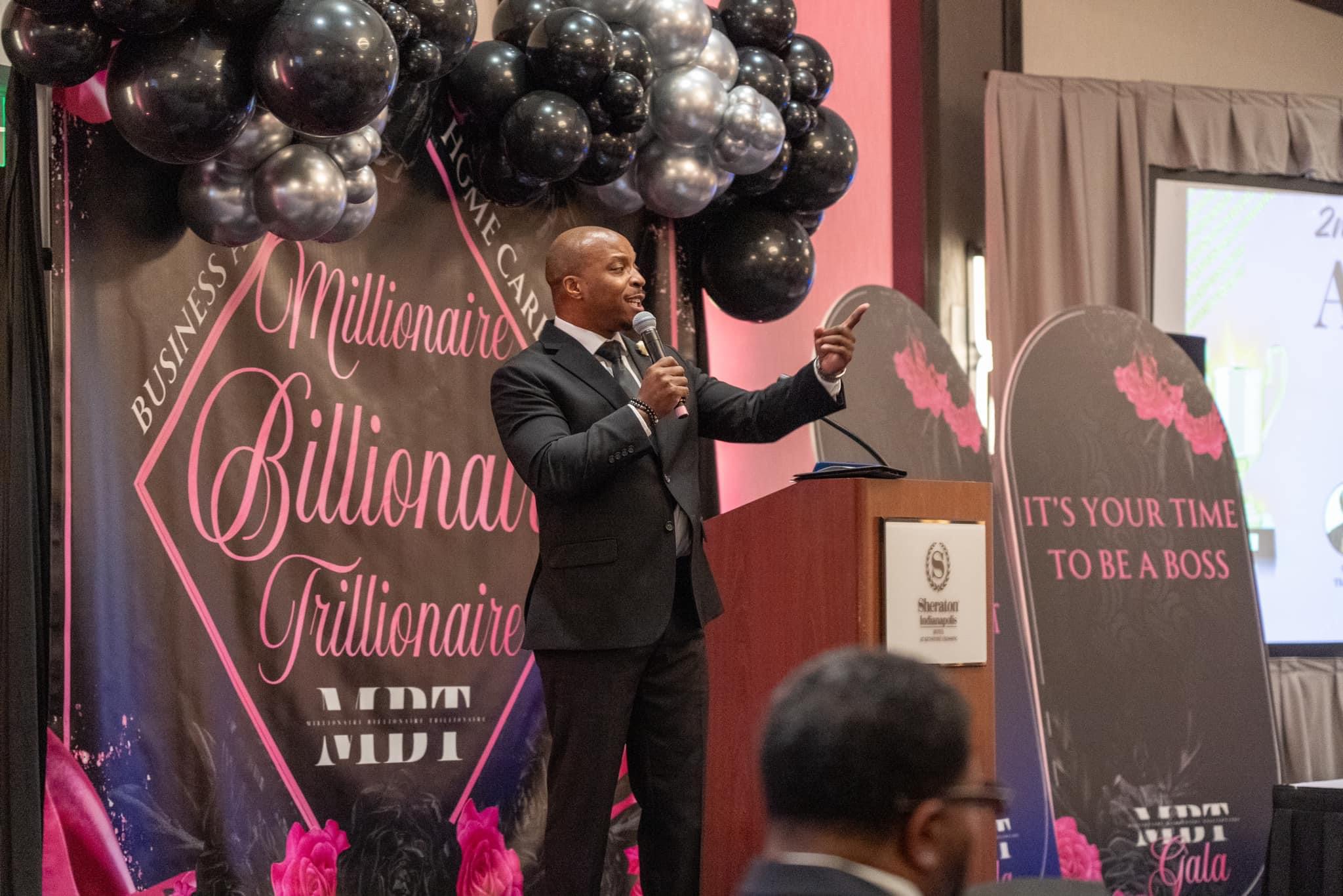 Man speaking at a podium during an event with balloons and banners, one banner reads 'It's your time to be a boss'.