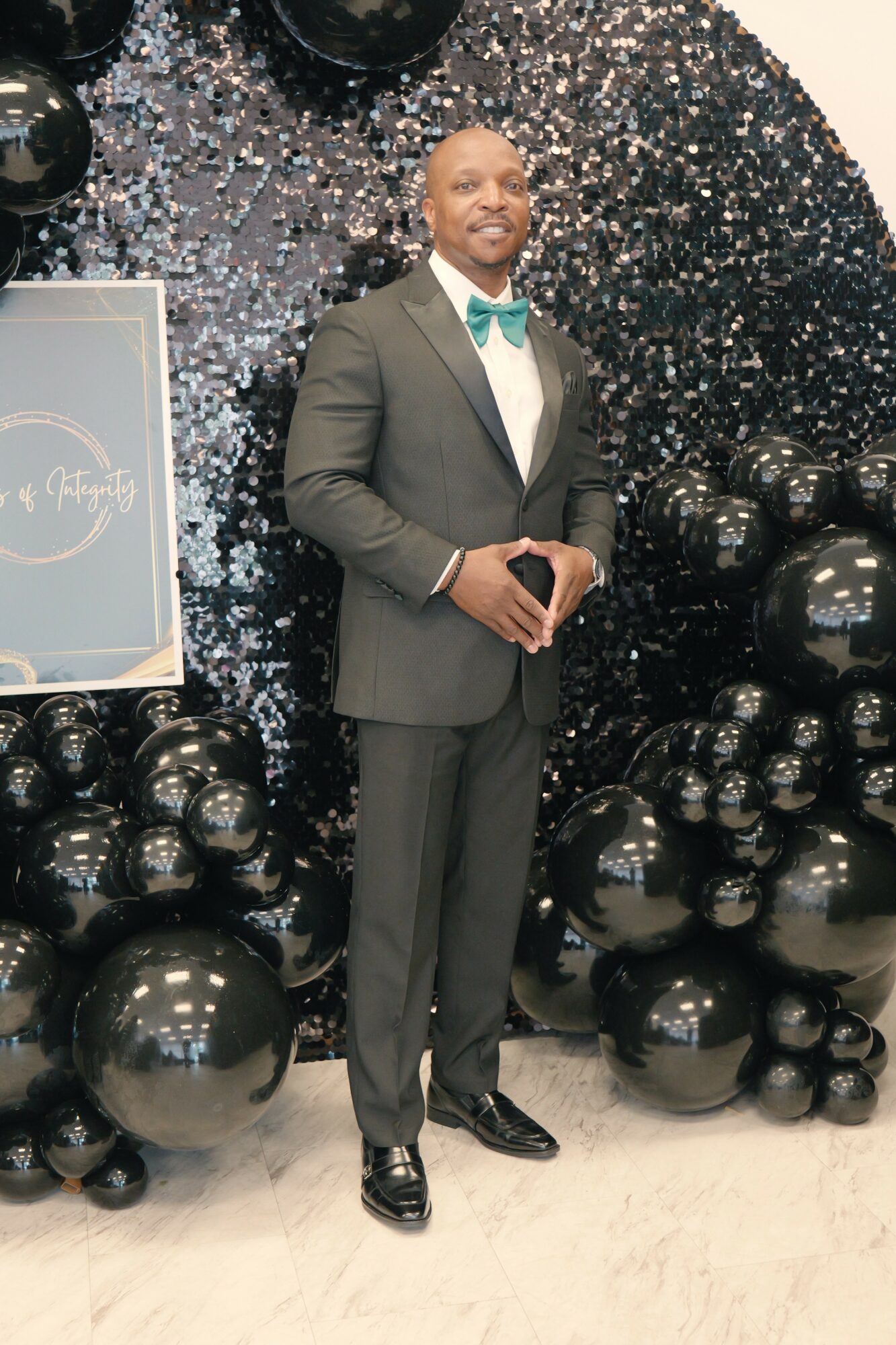 Man in a suit with a bow tie standing in front of a black and silver balloon backdrop, holding a phone.