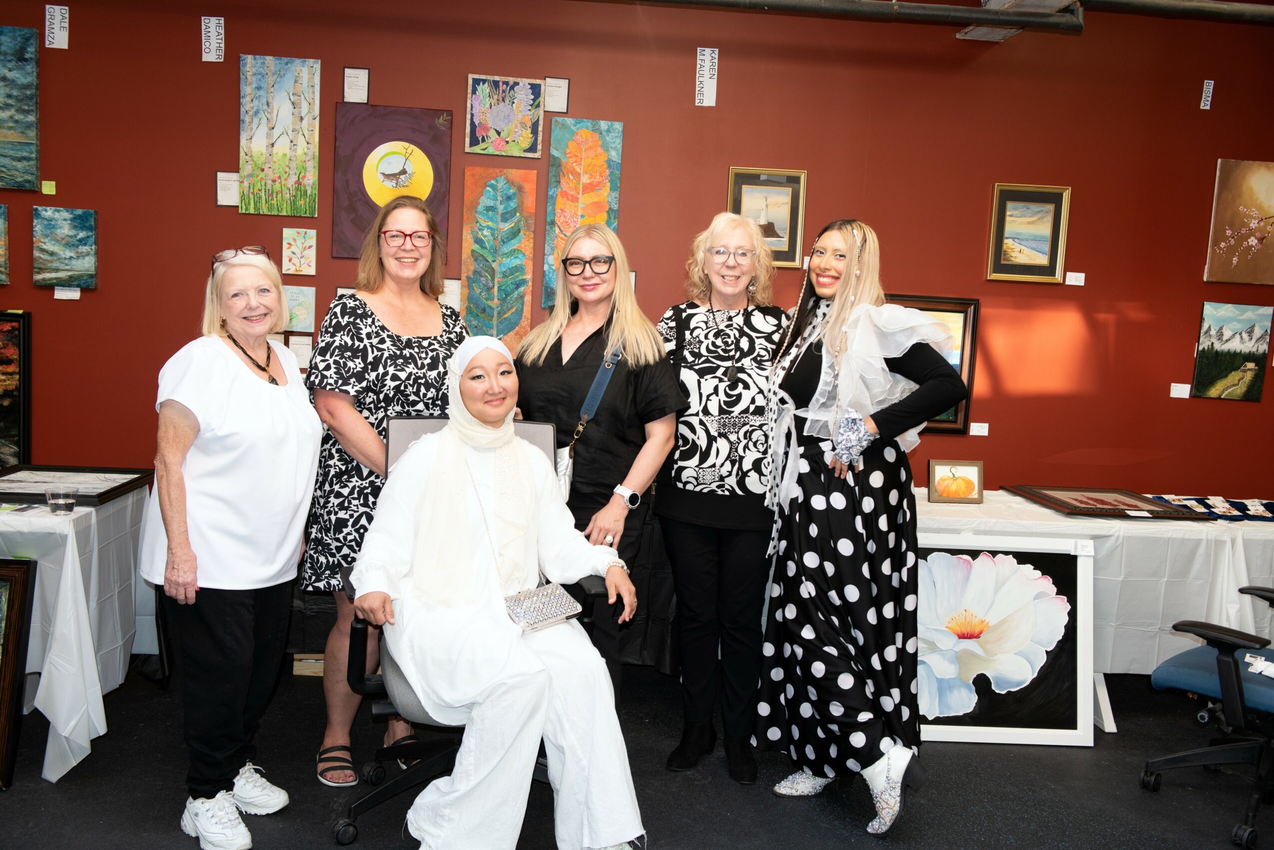 Six women standing and sitting in an art gallery with colorful paintings on the wall, smiling and posing for the photo.