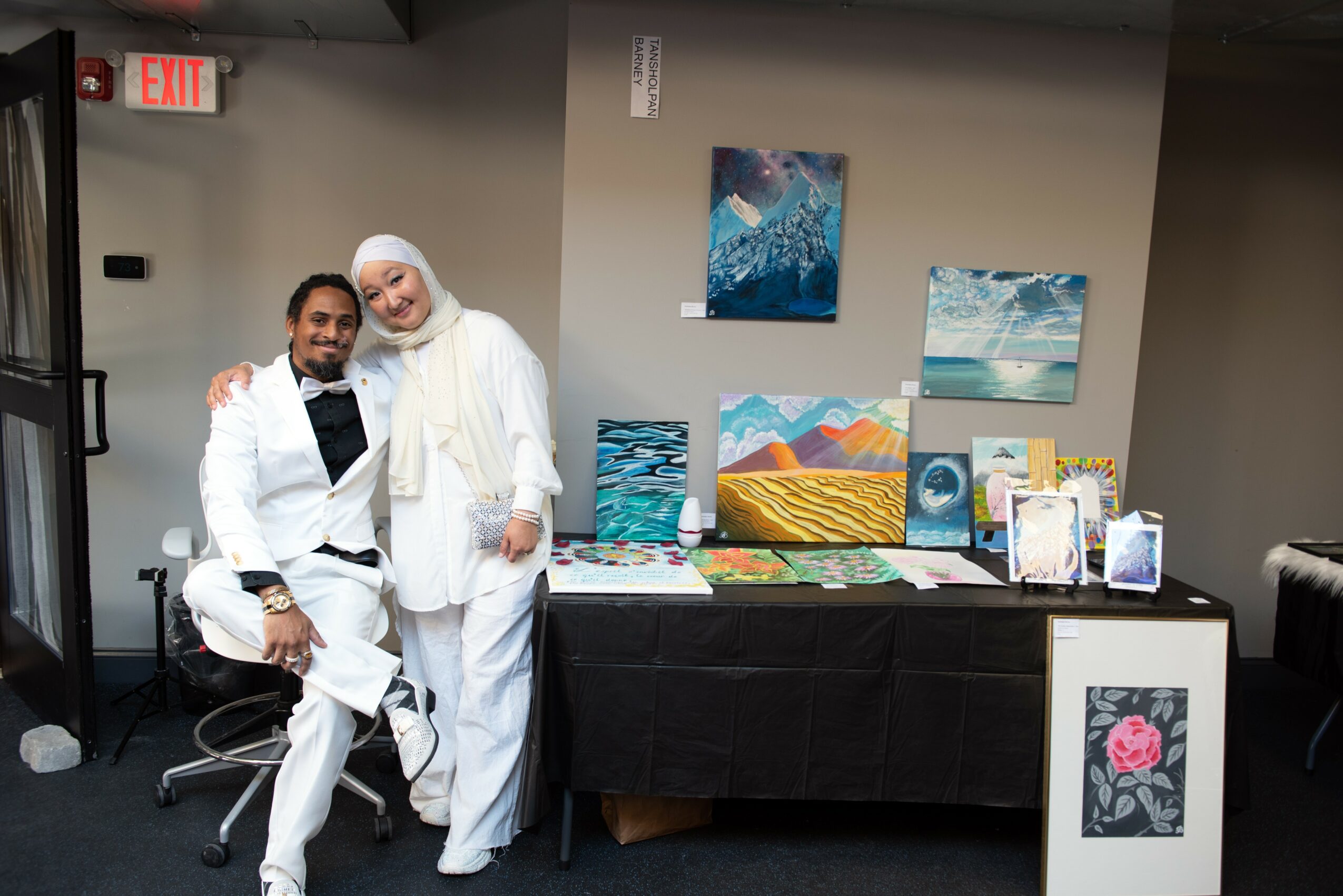 Two people in white outfits pose together in an art gallery with paintings on the wall behind them.