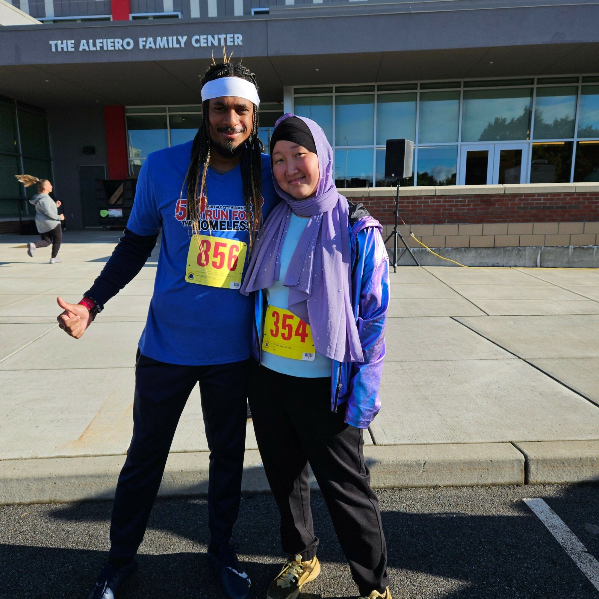 Two women standing outside a building with a sign reading 'The Allero Family Center.' They wear race bibs, one with '354.'
