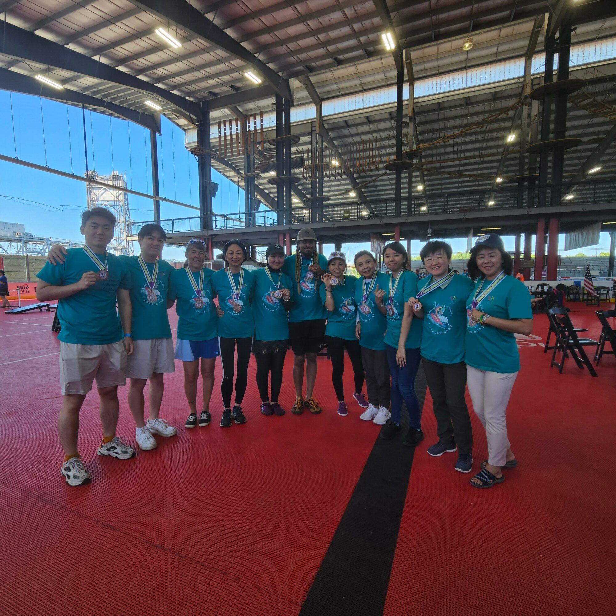 Group of ten people standing indoors, wearing matching teal shirts, smiling, with a large window and industrial ceiling in the background.