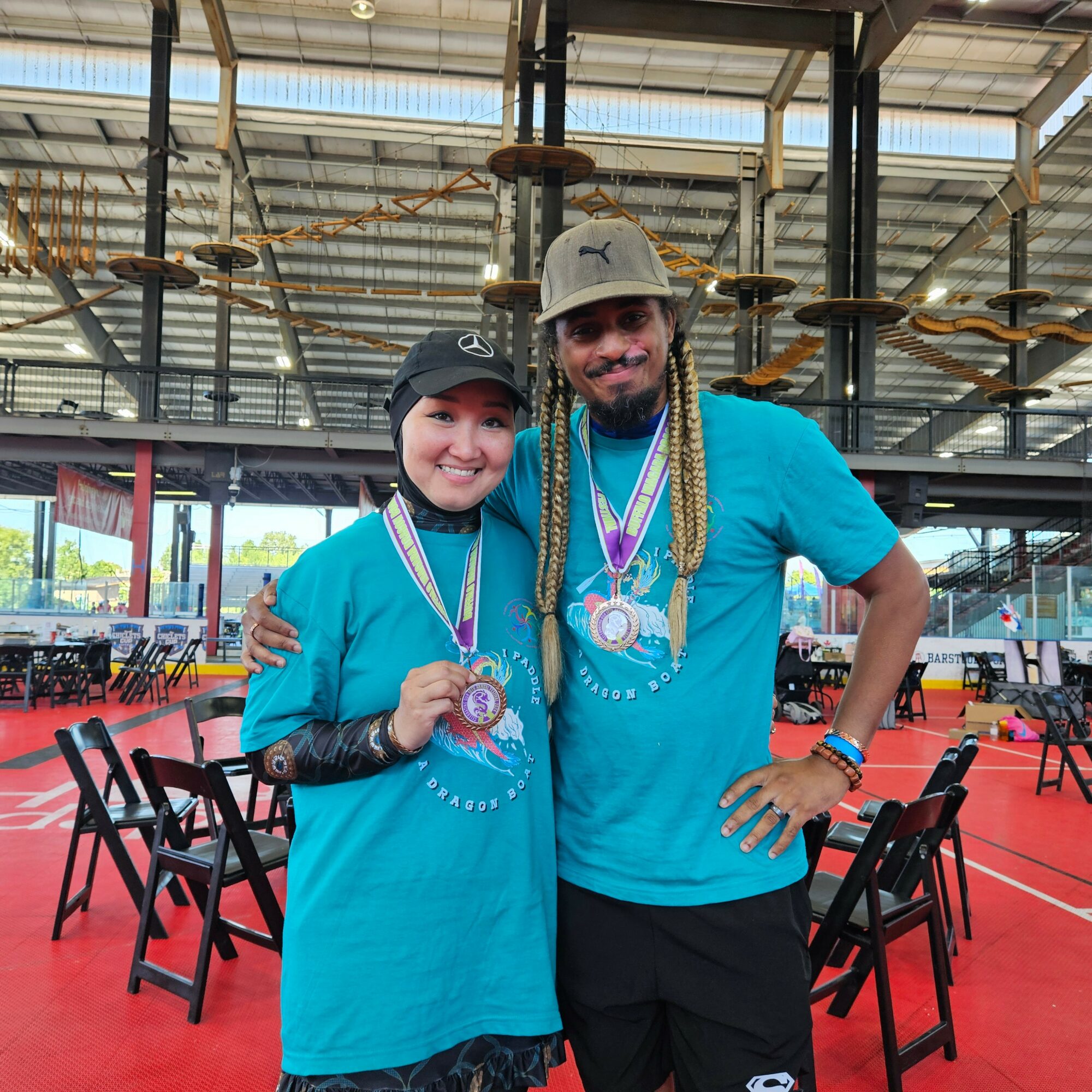 Two people wearing blue shirts and medals, standing indoors with chairs and a high ceiling in the background.