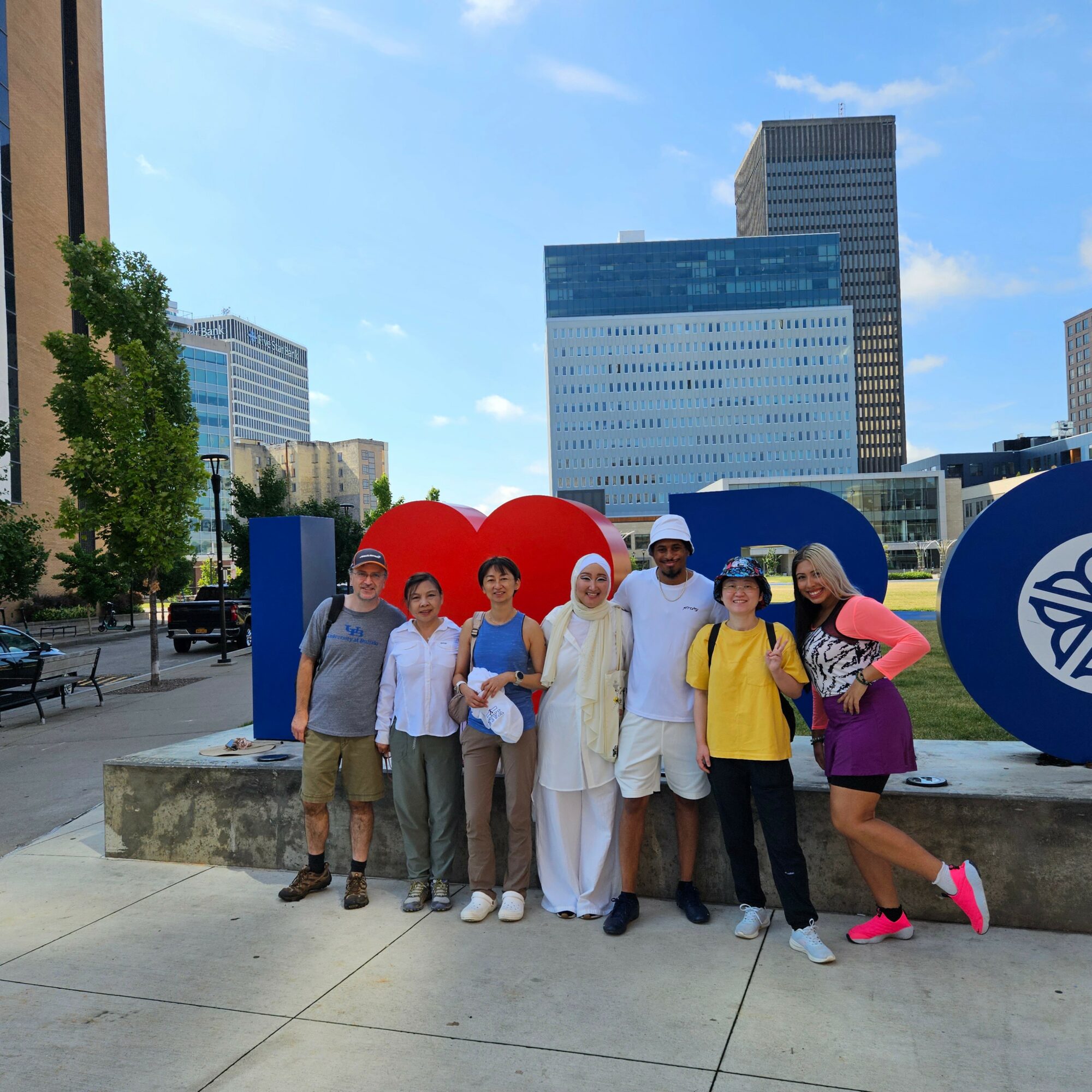 Group of eight people standing outdoors in front of large colorful letters and symbols, with city buildings in the background.
