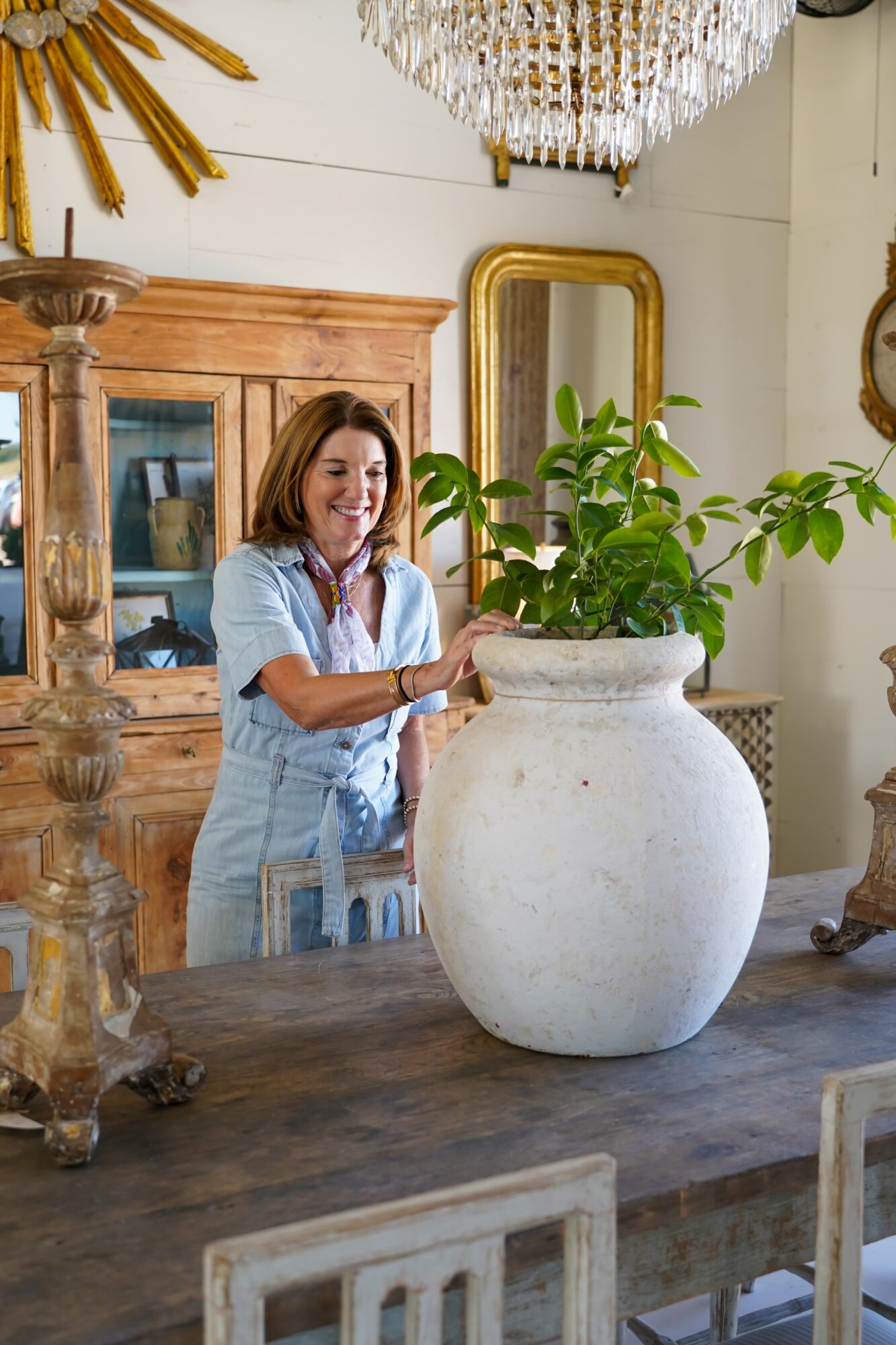 Woman with shoulder-length hair smiling, standing behind a large potted plant on a wooden table in a decorated room.