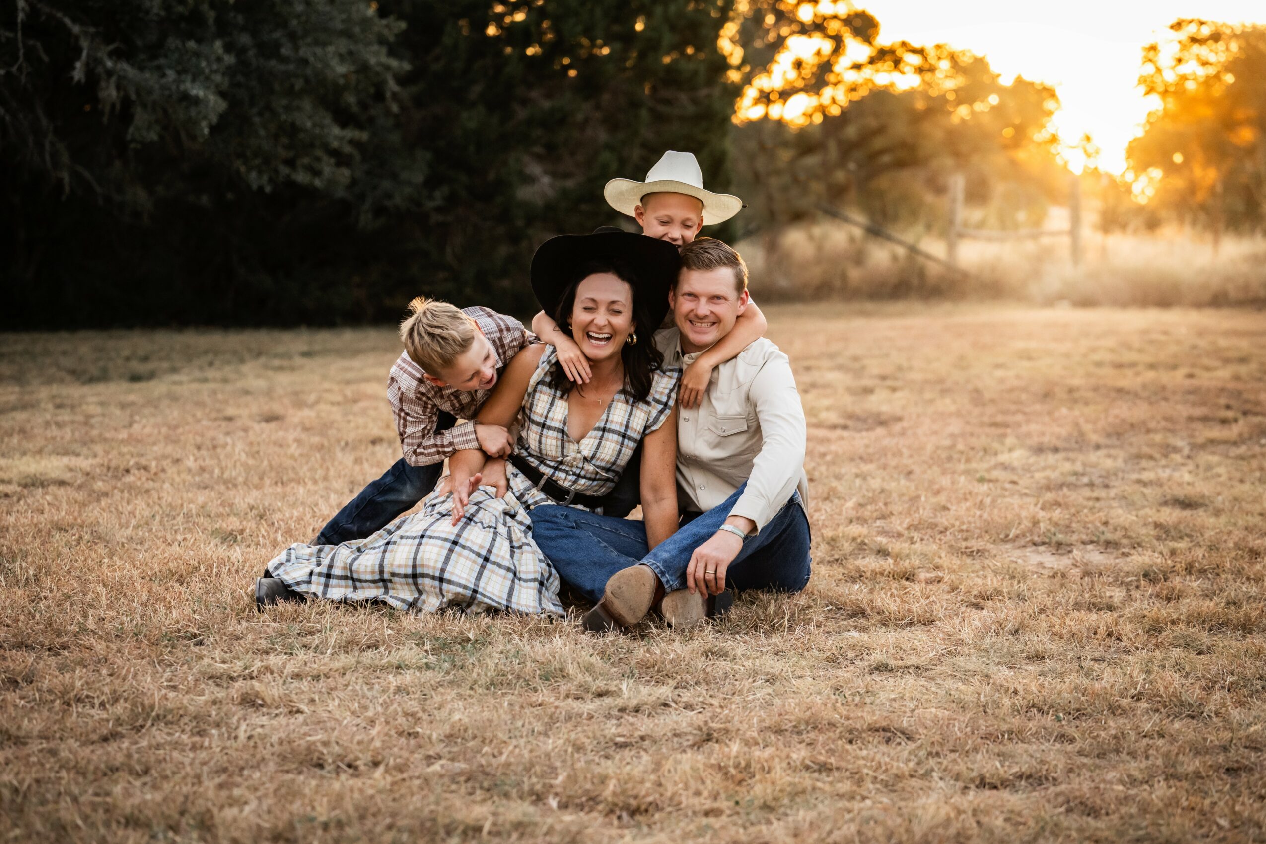 Family of four sitting on grass in a field during sunset, smiling and hugging, with trees in the background.