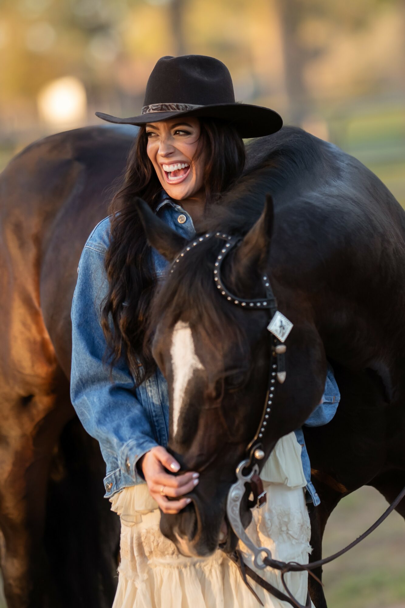 Woman smiling, wearing a black hat and denim jacket, holding a black horse with a decorative bridle outdoors.