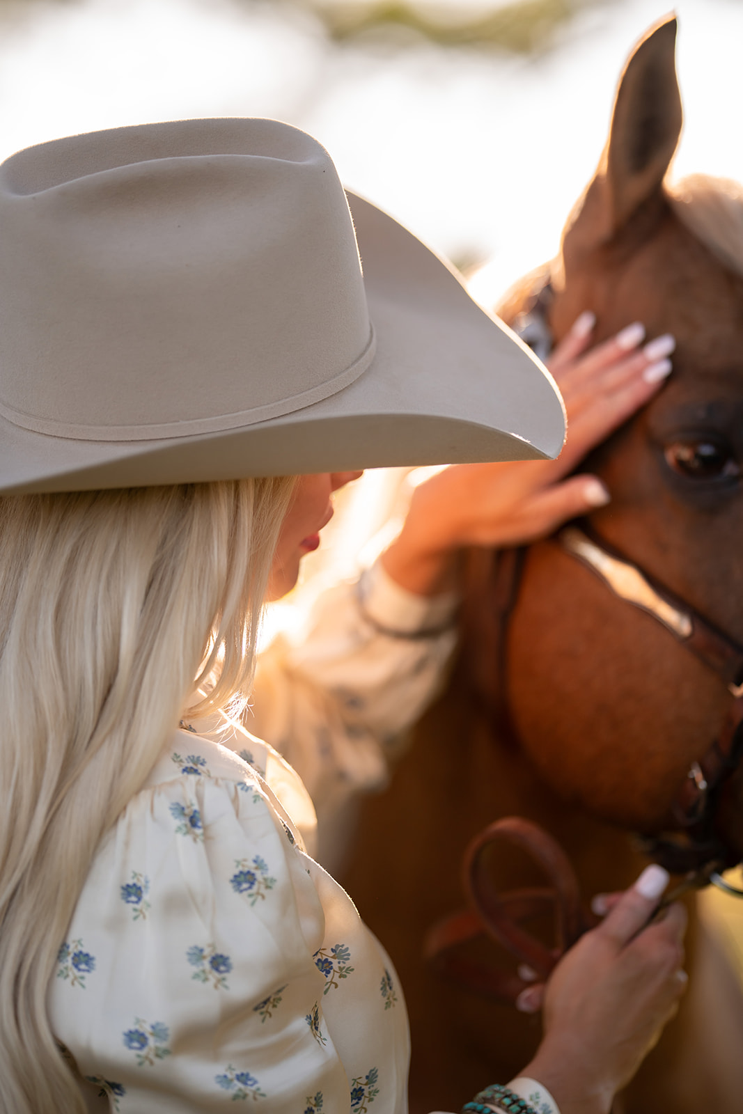 Person wearing a large white hat and floral blouse gently touches a brown horse's face.