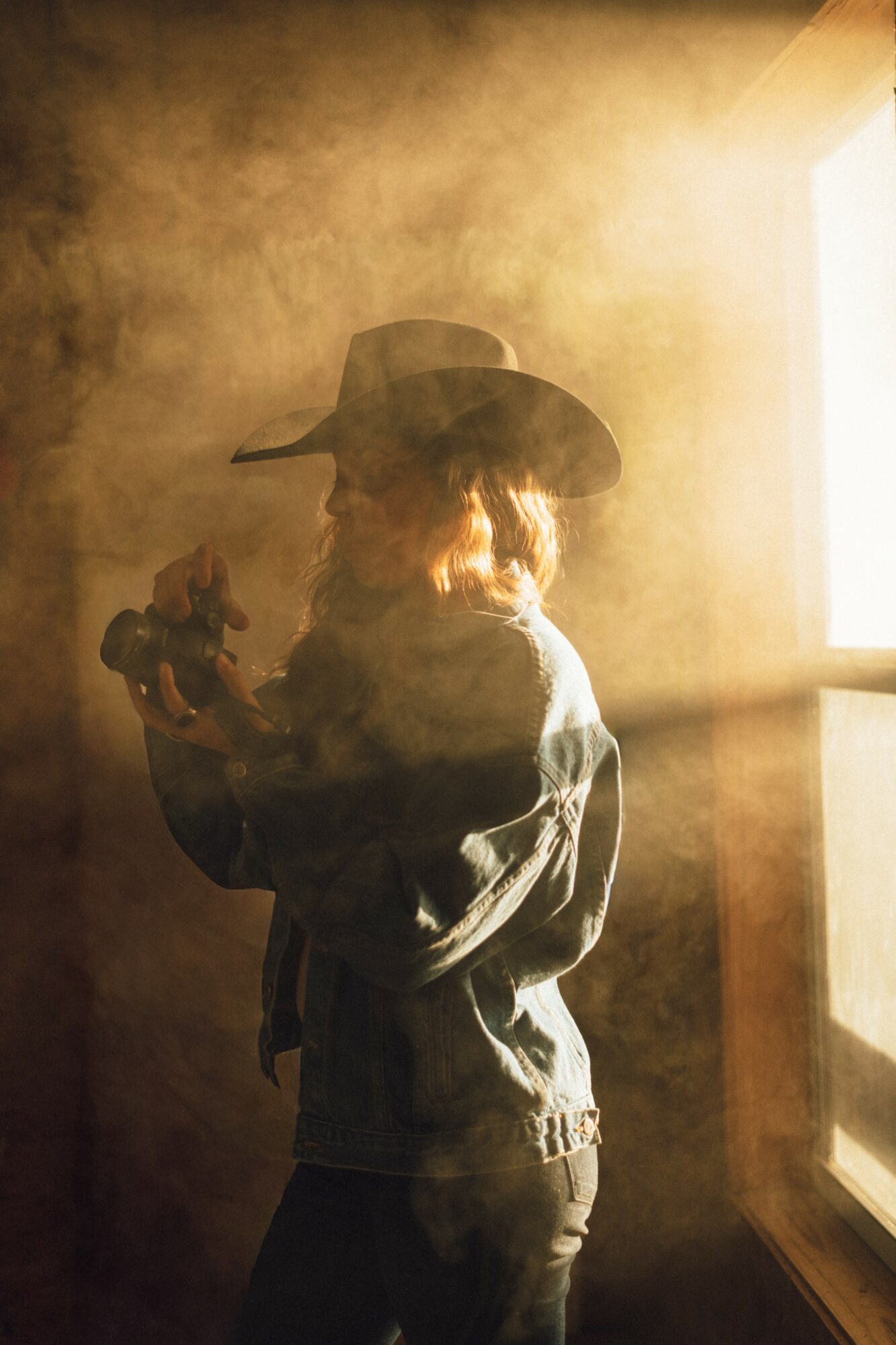Child wearing a hat and jacket, standing in a smoky room near a window, holding an object in hand.