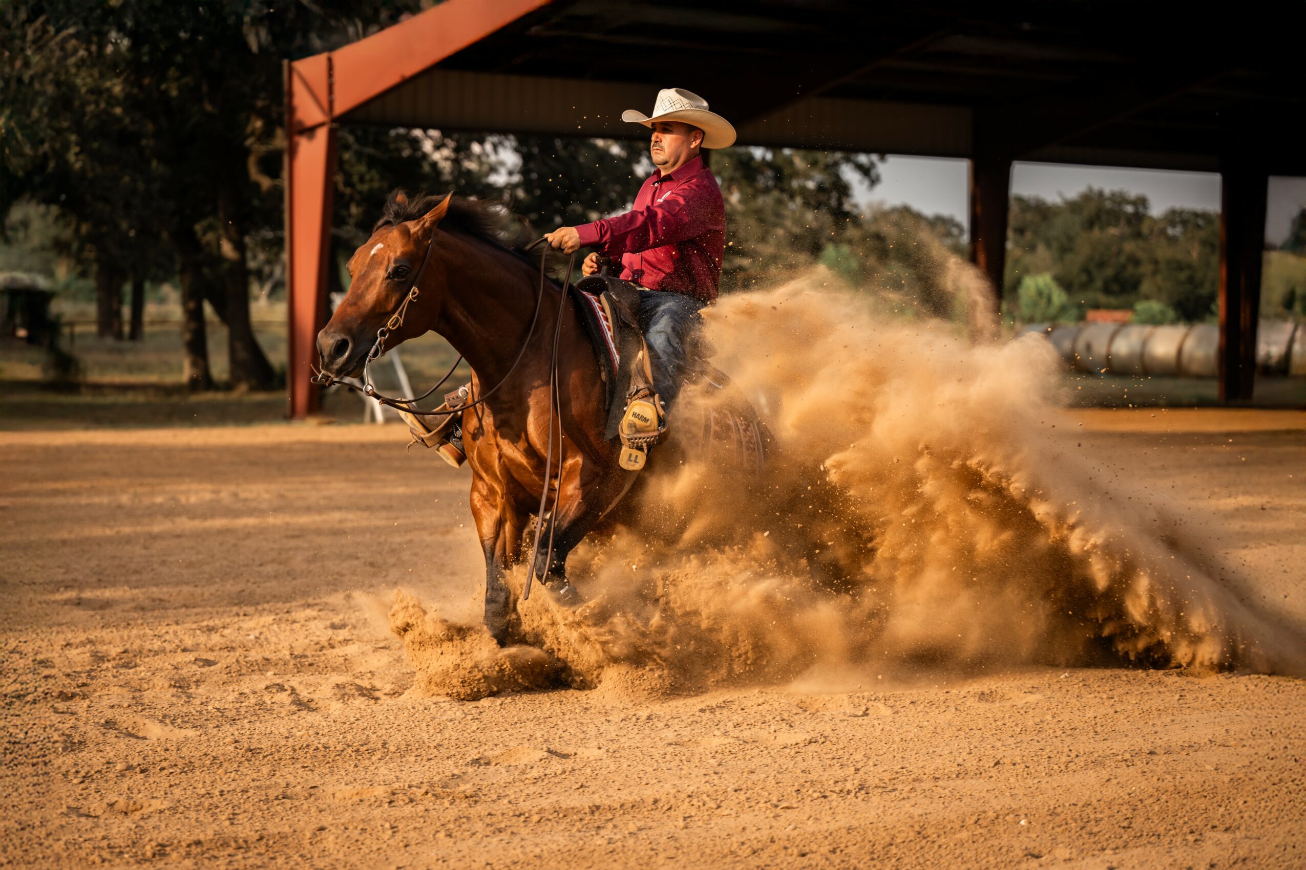 Person riding a horse kicks up dust in an outdoor arena, wearing a cowboy hat and red shirt.