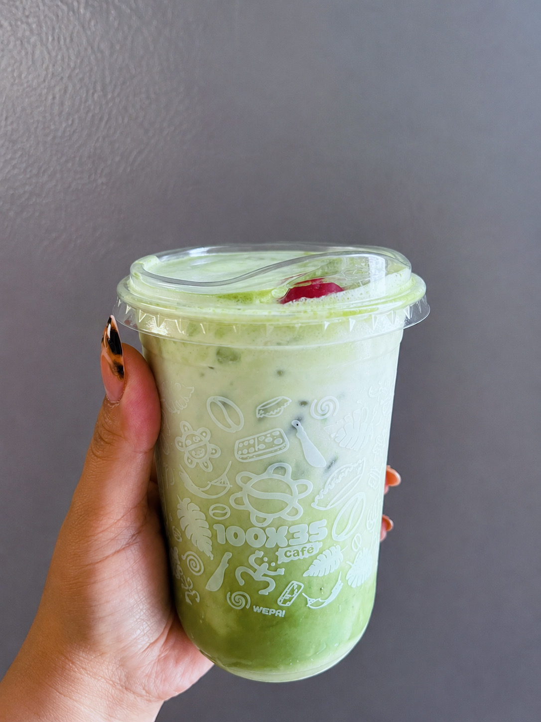 Hand holding a green beverage in a clear plastic cup with a lid, against a gray background.