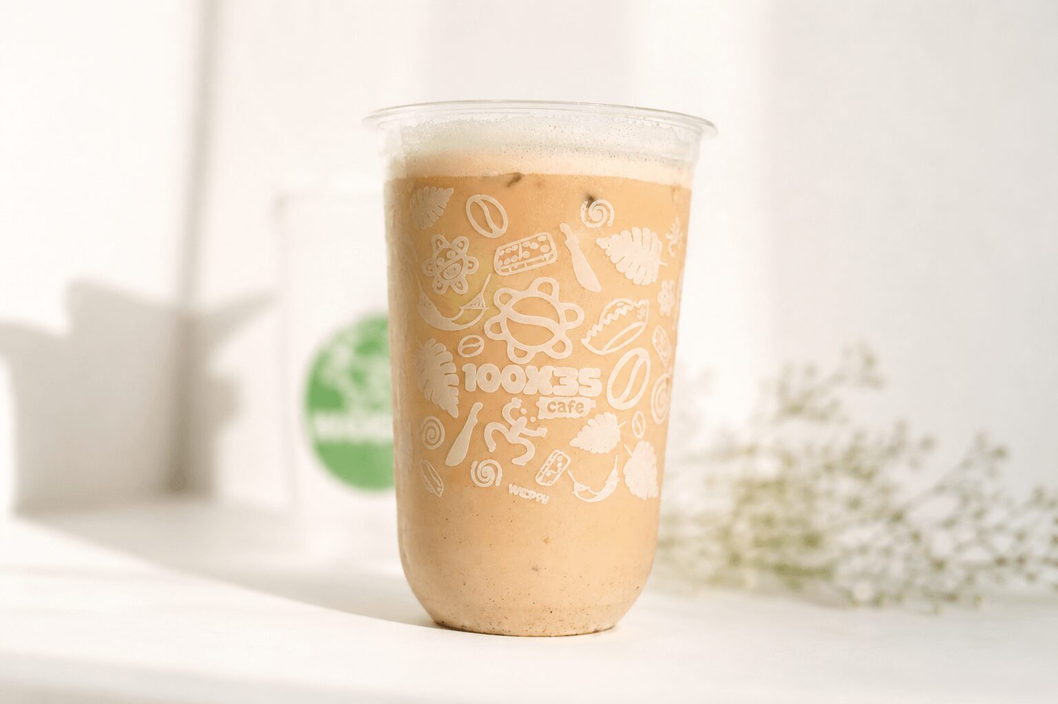 A clear plastic cup with a beige-colored beverage, decorated with white illustrations of coffee beans and cups, on a white surface.