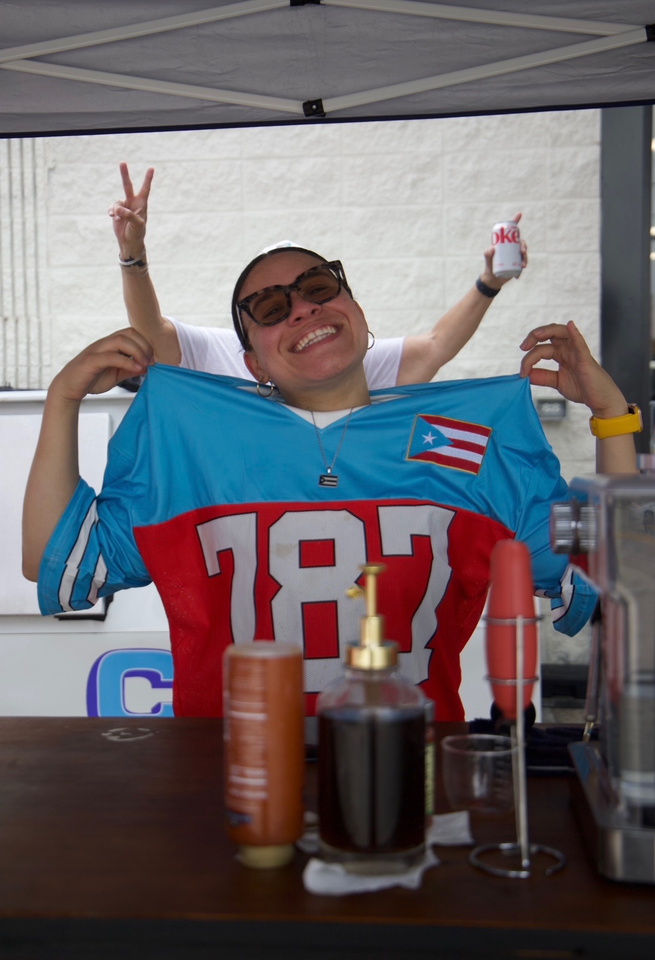 Person smiling, wearing sunglasses and a sports jersey, holding a flag, making a peace sign, with a tent and drinks in front.
