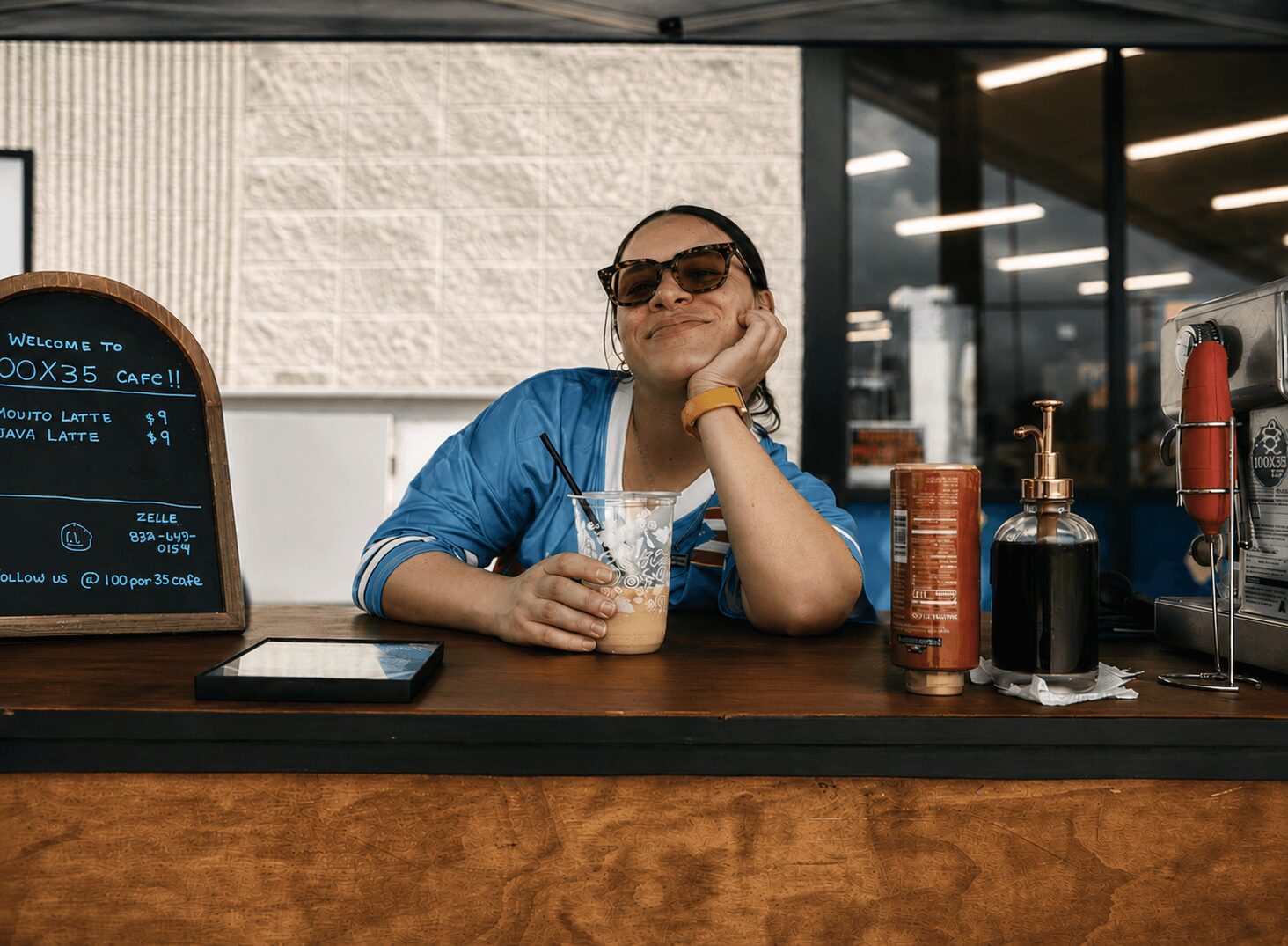 Woman sitting at a counter with sunglasses, smiling, holding a drink, in a cafe or restaurant setting.