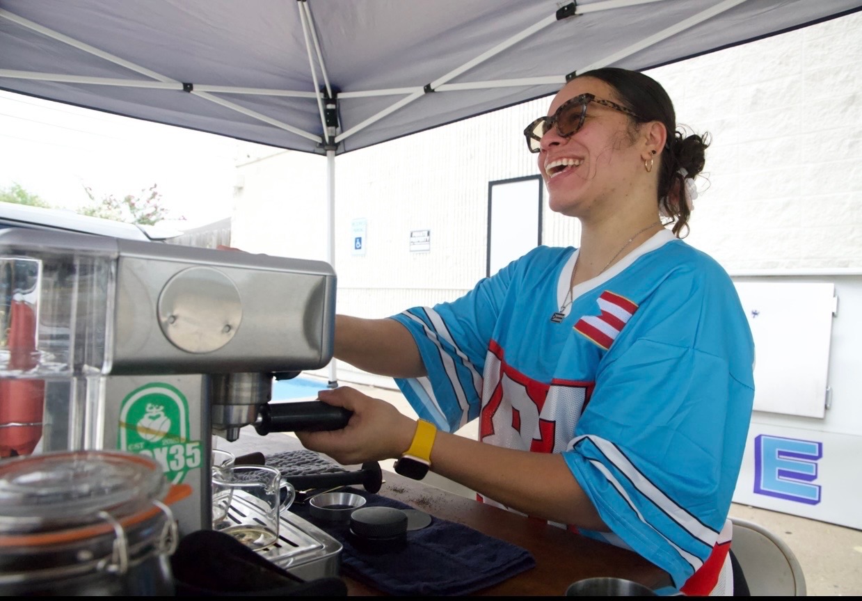 Woman smiling behind a coffee machine under a canopy, wearing a blue sports jersey and glasses.