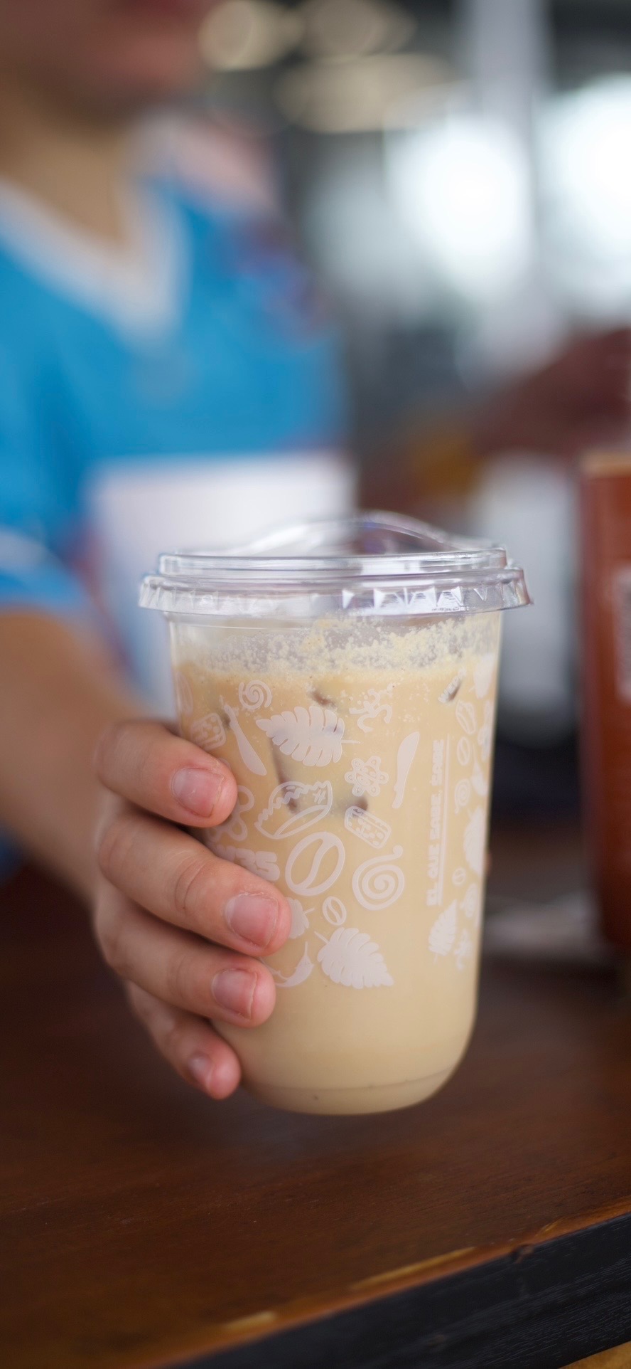 Person holding a plastic cup with a creamy beverage, blurred background, wooden surface, and person in blue shirt.