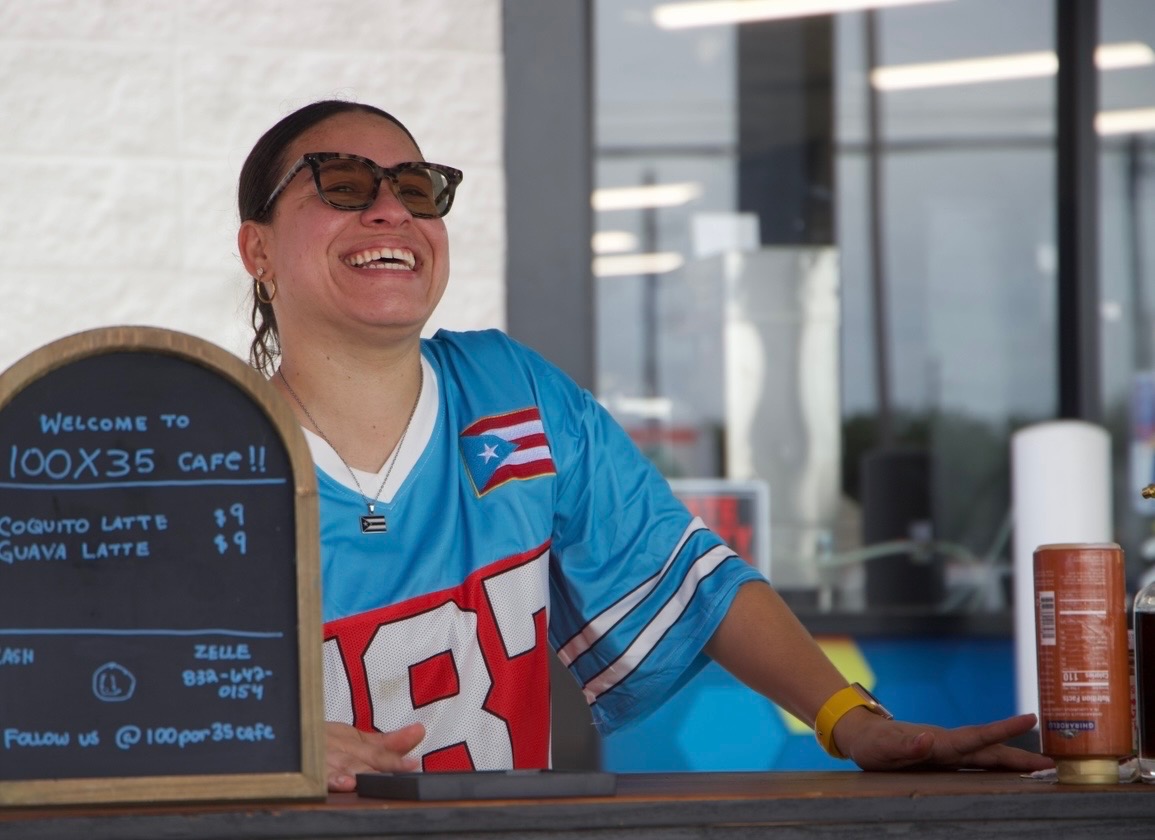 Woman smiling behind a counter with a chalkboard menu, wearing sunglasses and a blue sports jersey.