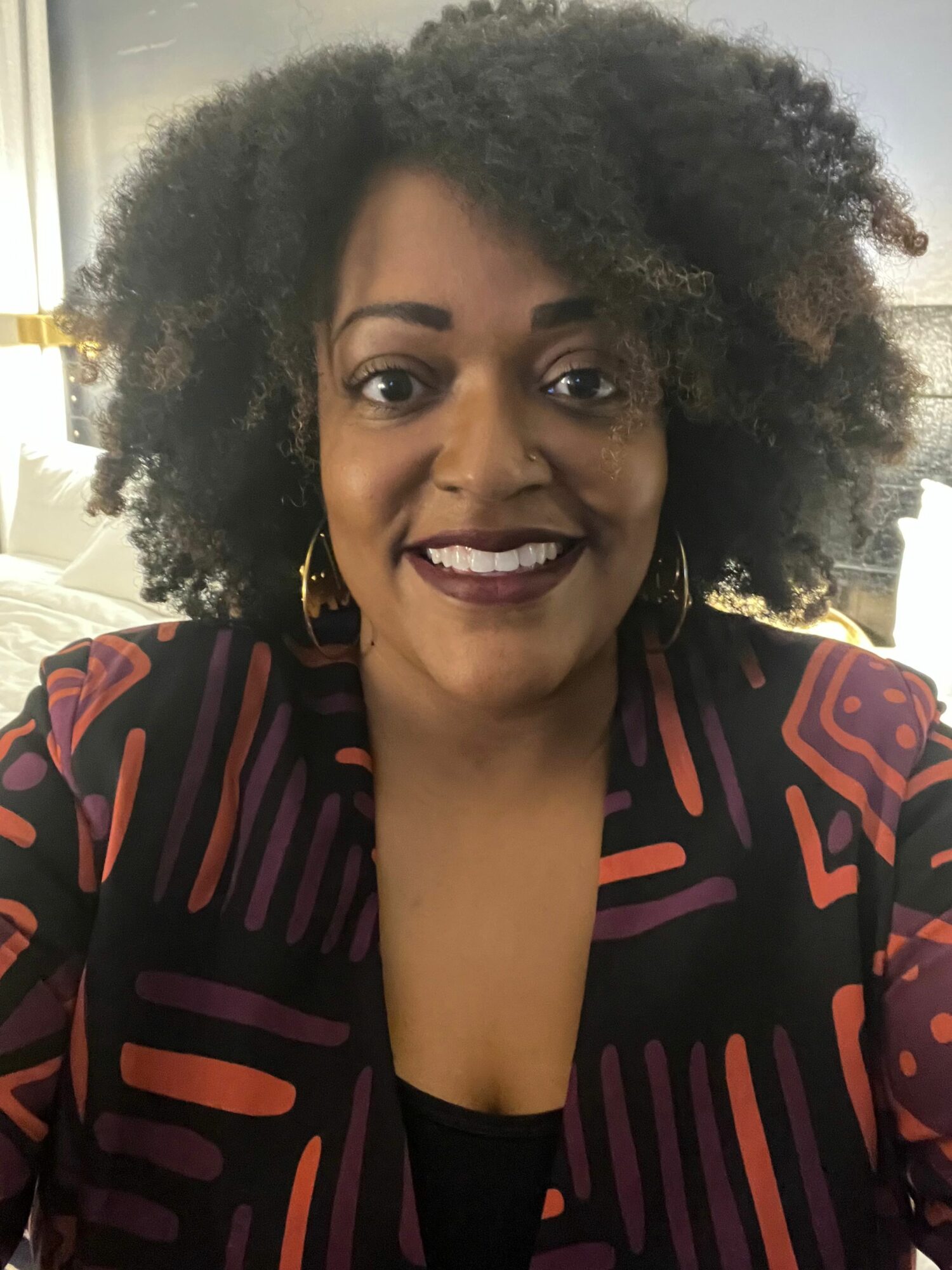 Woman with curly hair smiling, wearing a patterned black and red top, indoors with a blurred background.