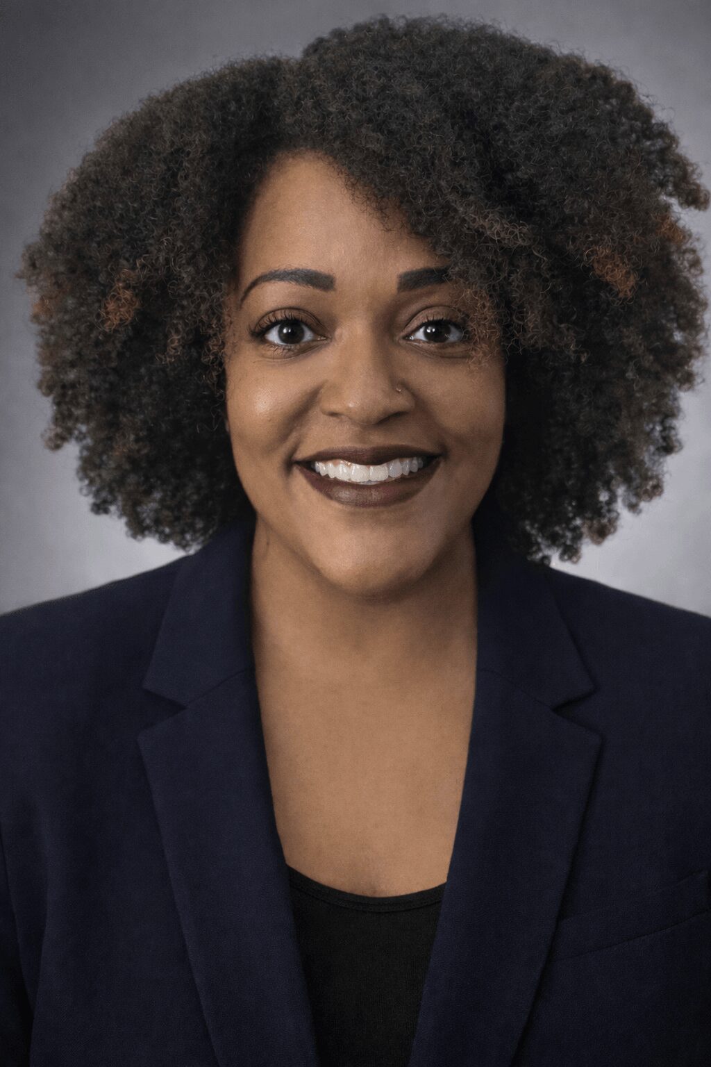 Portrait of a woman with curly hair, smiling, wearing a dark blazer and top, against a neutral background.